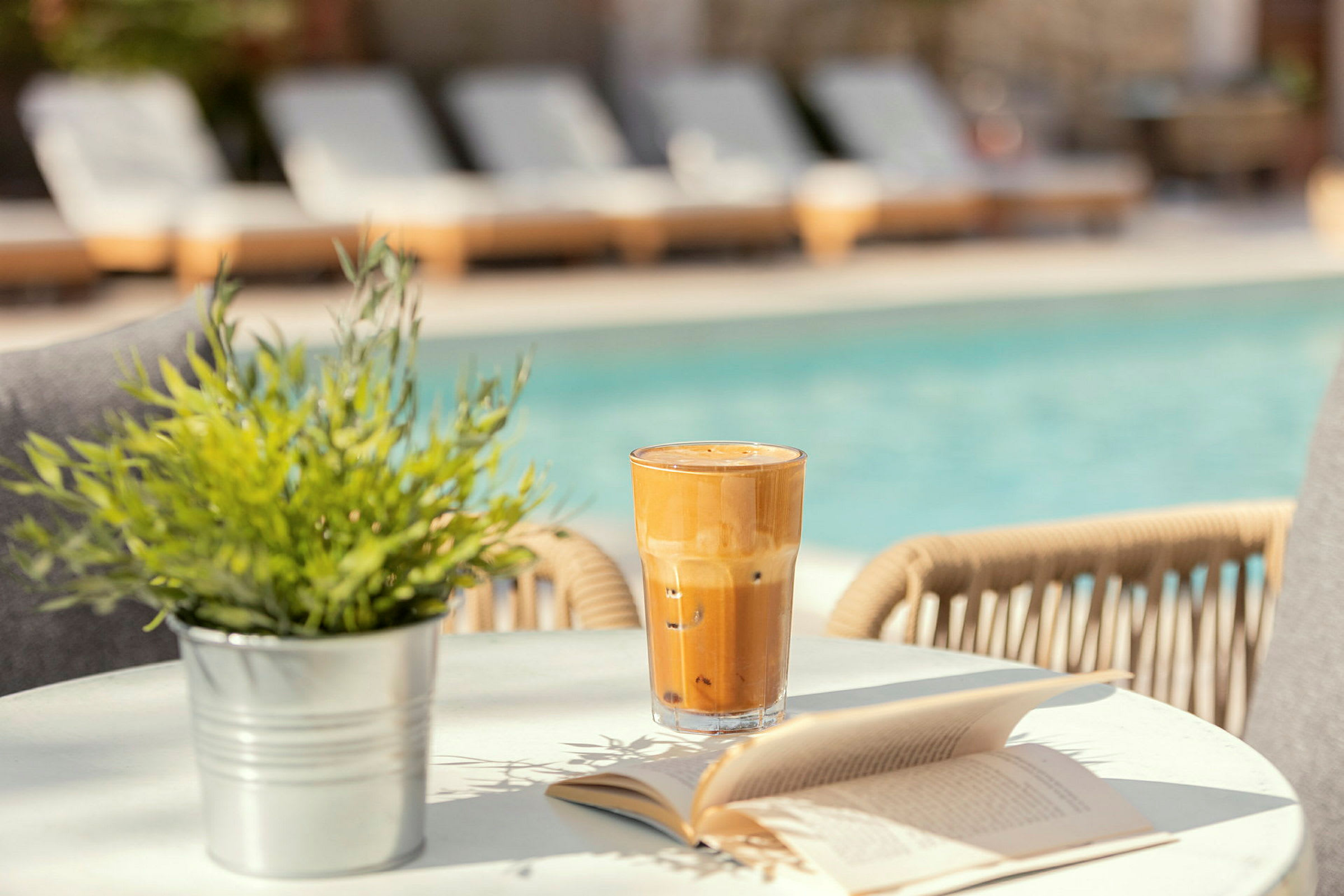 Ett glas iskaffe och en uppslagen bok på ett bord vid en pool. (AI-genererad text)