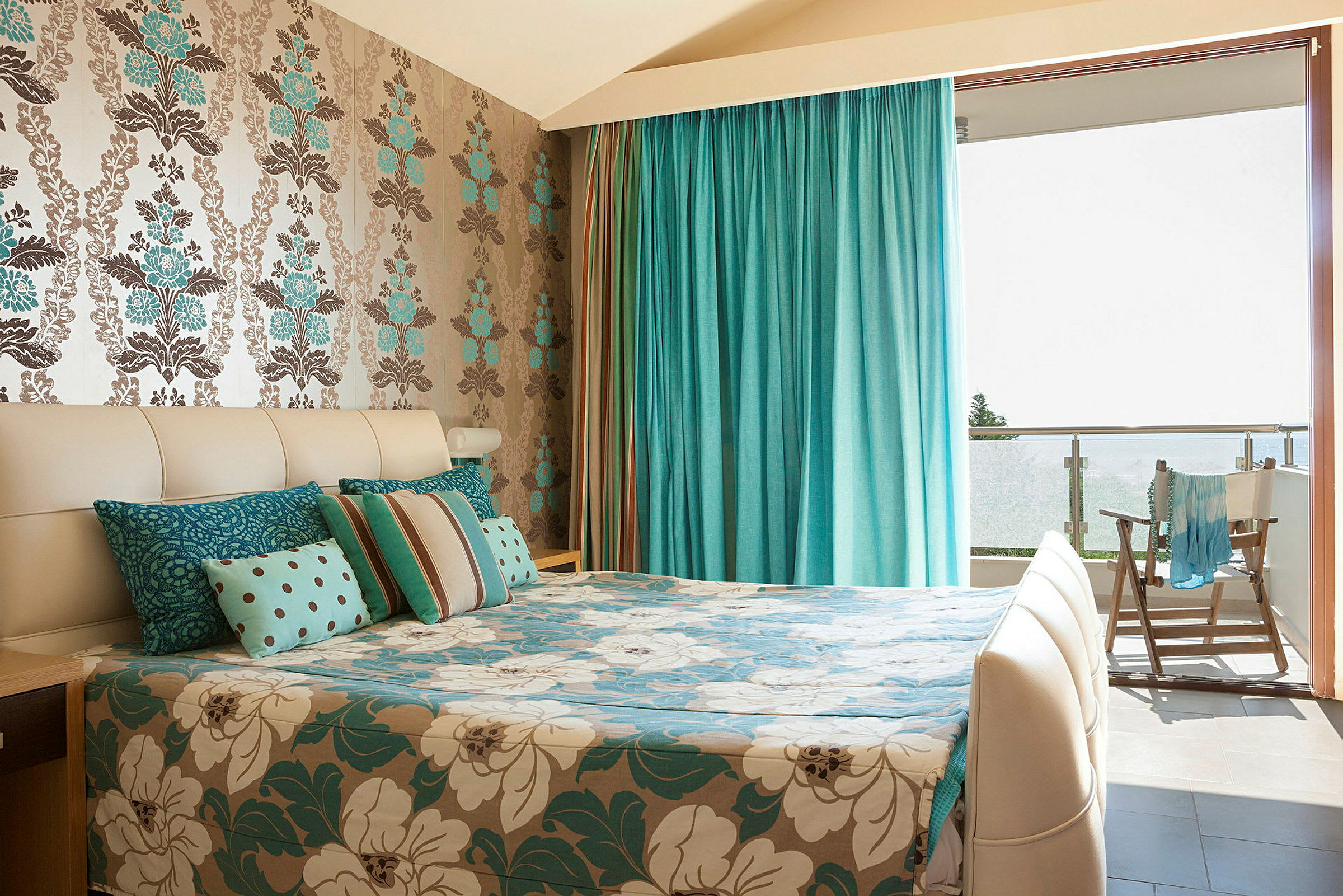 Ett rum med en säng dekorerad med blå och vita blommiga mönster, en fondvägg med blommotiv och draperier i turkos som leder till en balkong. (AI-genererad text)