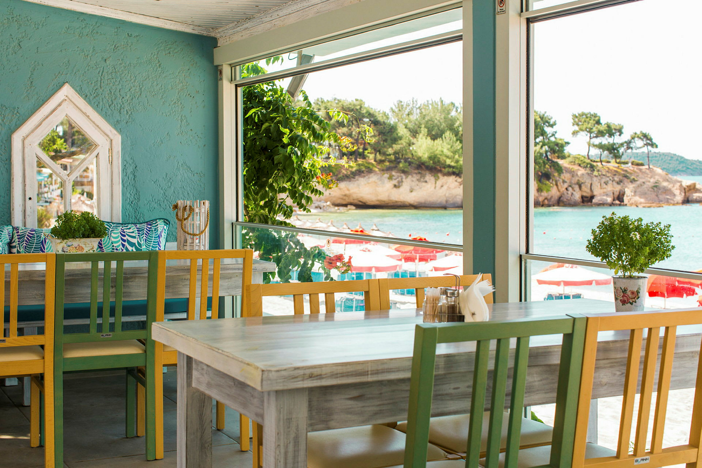 En ljus restaurang med färgade trästolar och bord, med utsikt över havet genom stora fönster. (AI-genererad text)