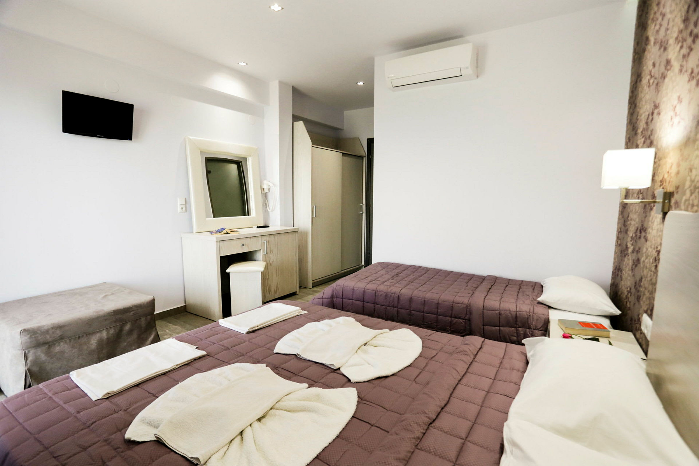 Ett hotellrum med två bäddar klädda med lila sängöverkast och vita handdukar. Rummet har en väggmonterad TV, ett skrivbord med spegel och ett luftkonditioneringsaggregat. (AI-genererad text)