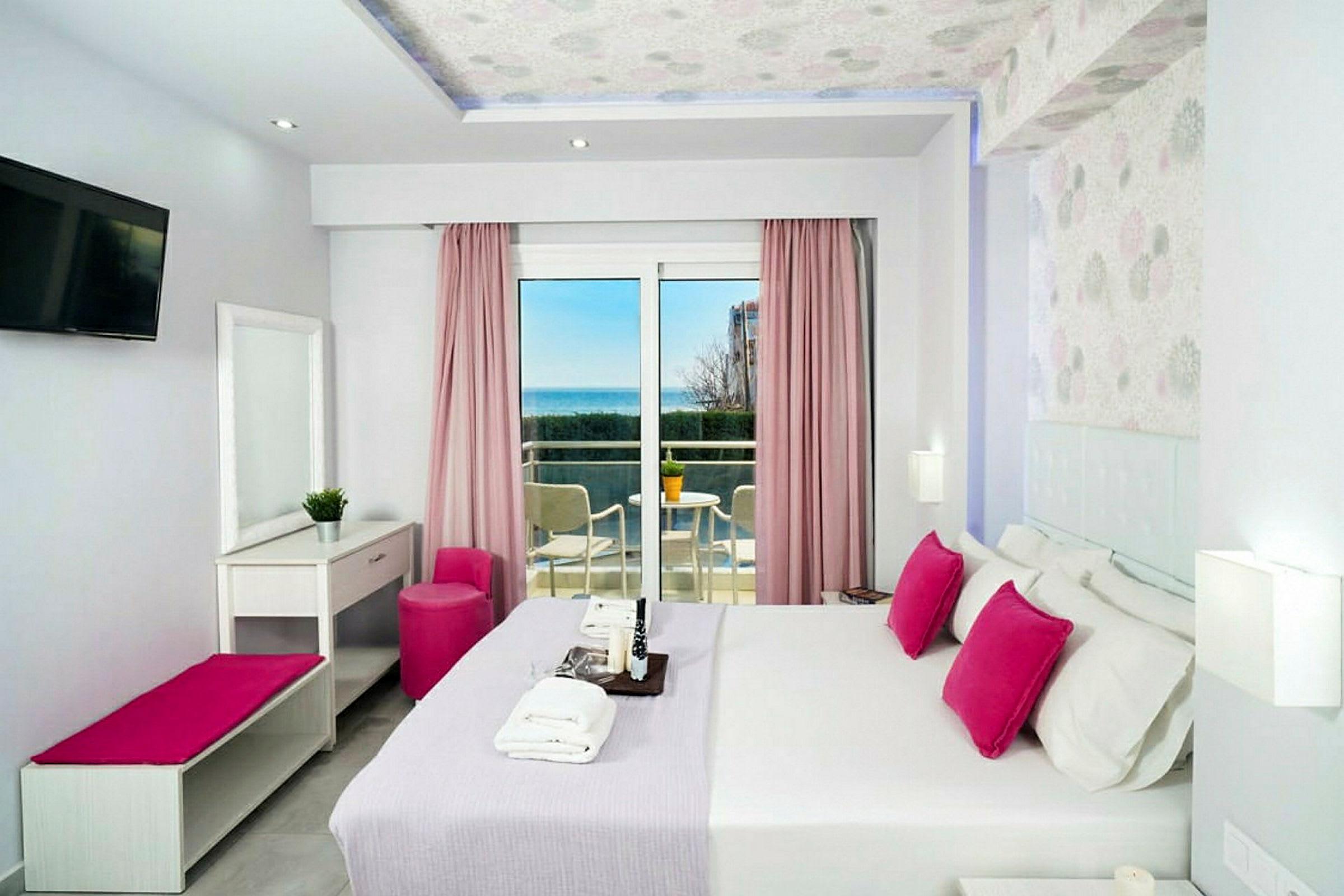 Ett hotellrum med ljus inredning, rosa detaljer och en balkong med utsikt över havet. (AI-genererad text)