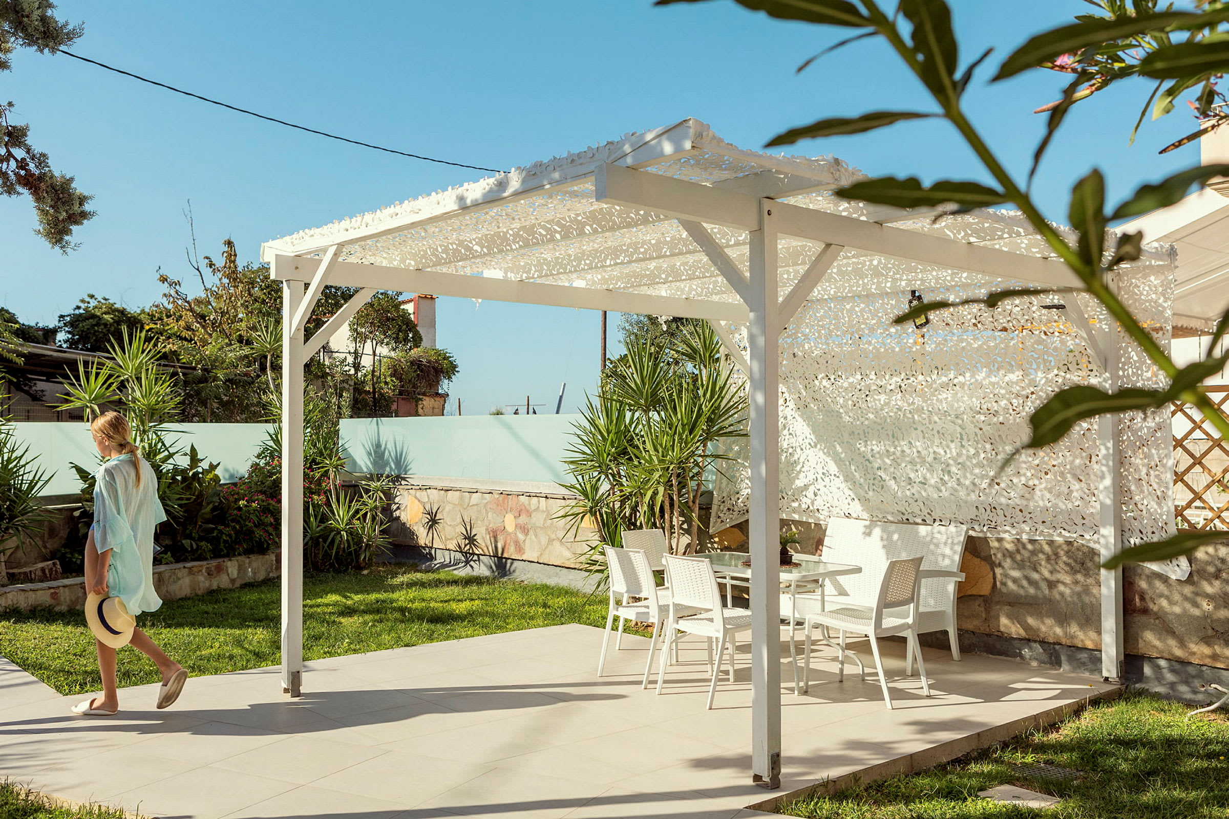 En person går intill en pergola med vita trädgårdsmöbler på en stenlagd yta, omgiven av grönska. (AI-genererad text)