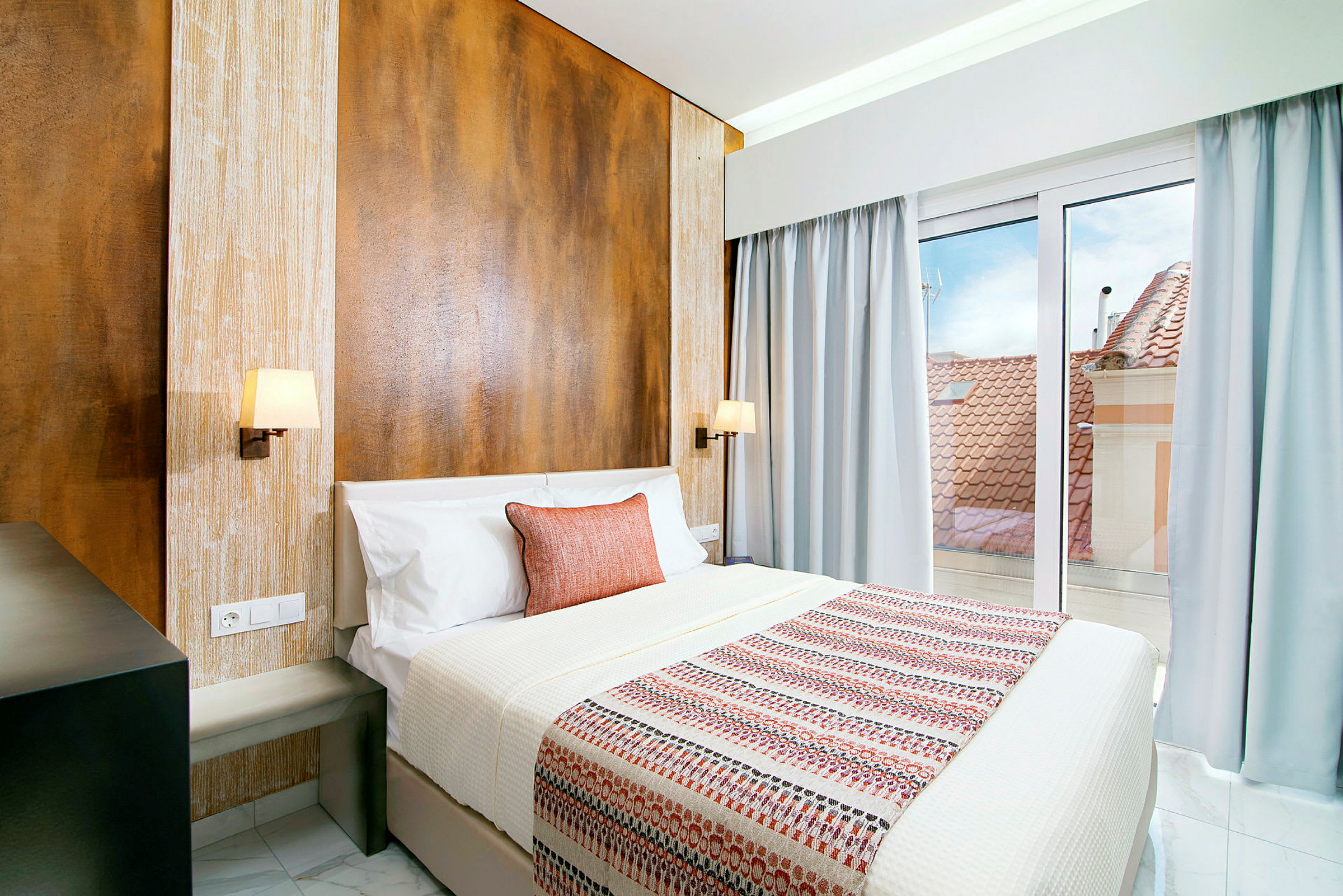 Hotellrum med en dubbelsäng, sängkläder i vitt och mönstrad bäddning. En kudde i orange ligger på sängen. Bakom sängen finns en trävägg med två vägglampor, och rummet har ett stort fönster med utsikt över tak och himmel. (AI-genererad text)