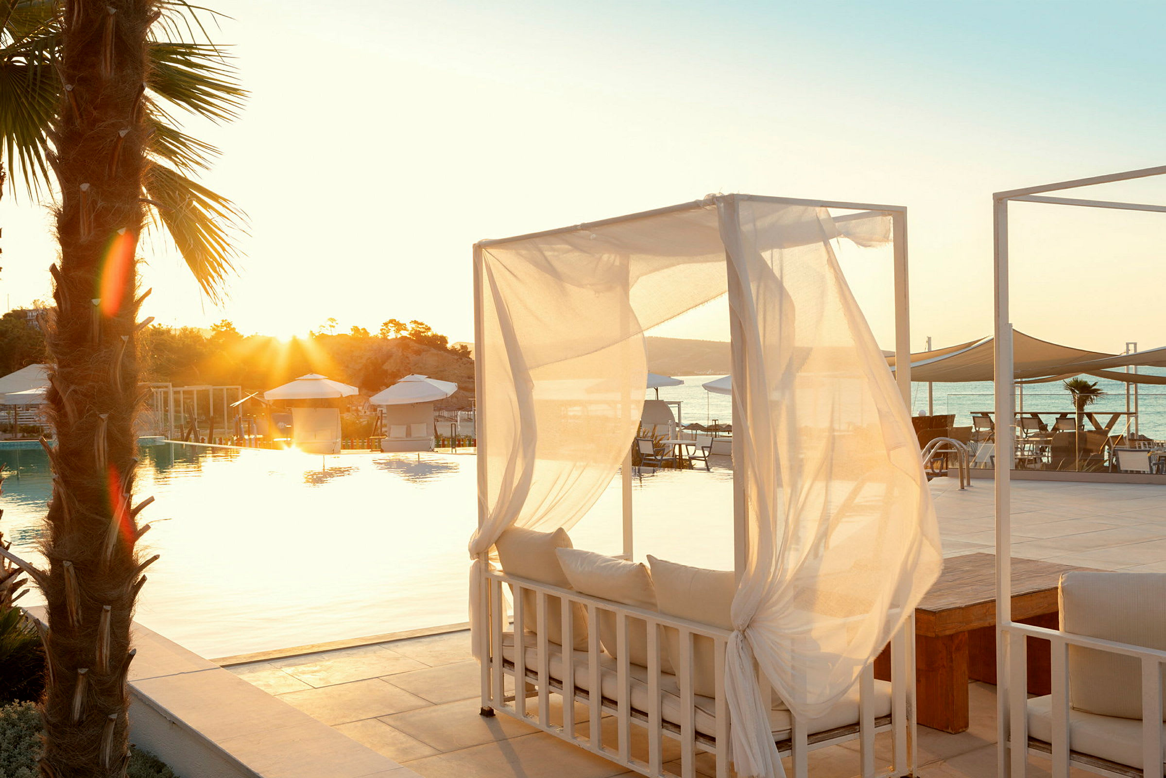 En pool med solsängar och parasoller vid solnedgång. En dagbädd med vita draperier i förgrunden. (AI-genererad text)
