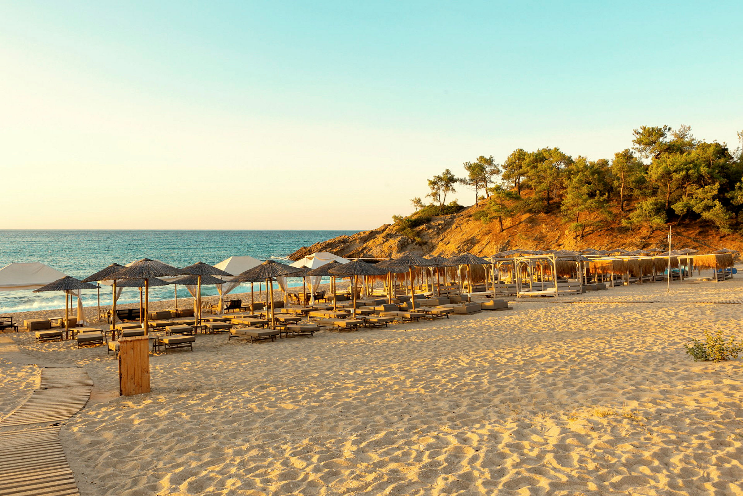En sandstrand med solstolar och parasoll, havet och skogbevuxen kulle i bakgrunden. (AI-genererad text)