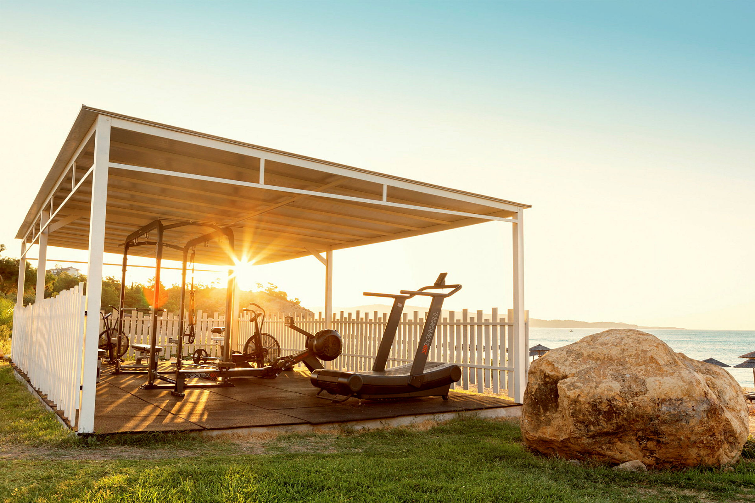 Ett utomhusgym under ett tak med träningsutrustning, beläget nära havet och med en stor sten i förgrunden. (AI-genererad text)