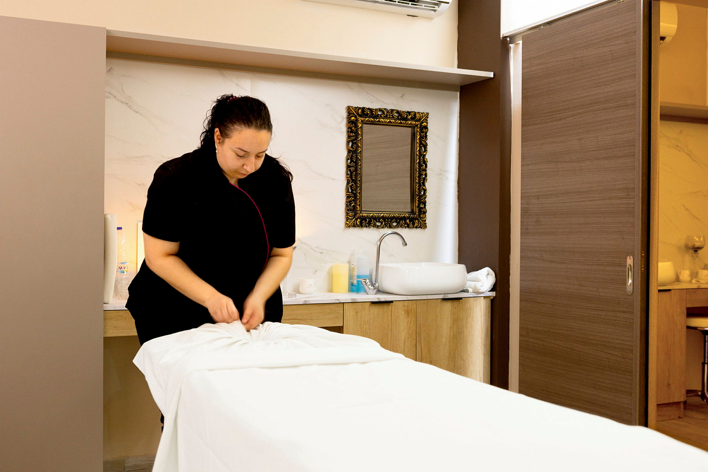 En person förbereder en massagebänk i ett spa eller behandlingsrum. I bakgrunden finns ett handfat och spegel. (AI-genererad text)