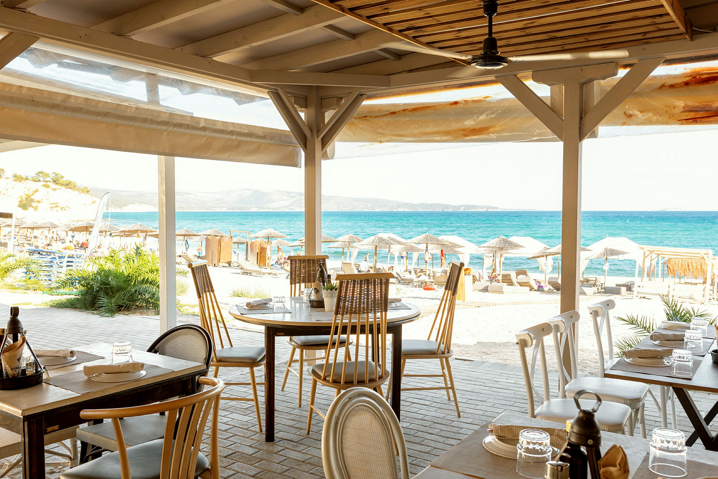 En restaurang med utsikt över stranden och havet, med uppställda bord och stolar under ett tak. (AI-genererad text)