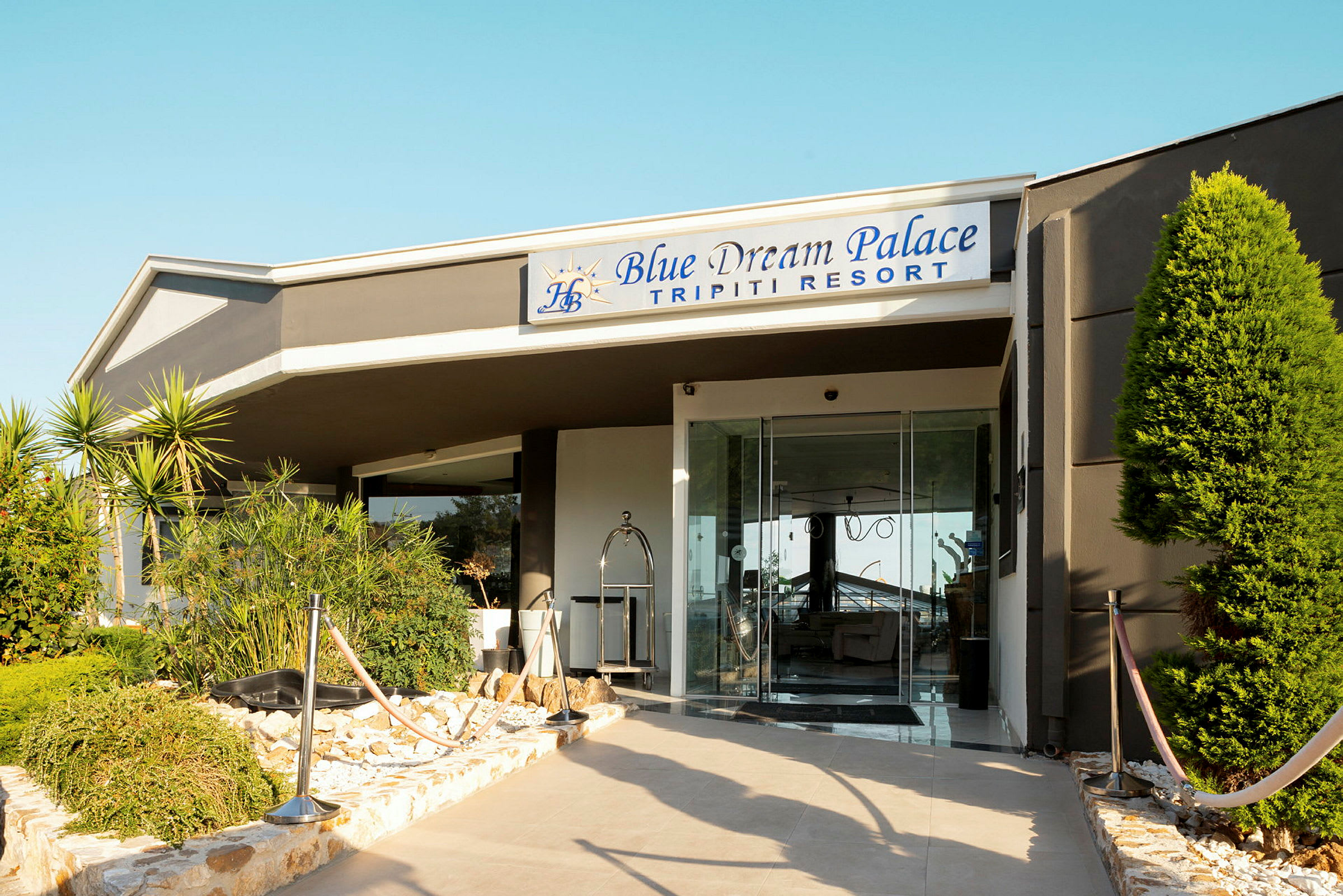 Ingång till ett hotell med skylt som lyder "Blue Dream Palace Tripiti Resort" och omgiven av gröna växter. (AI-genererad text)