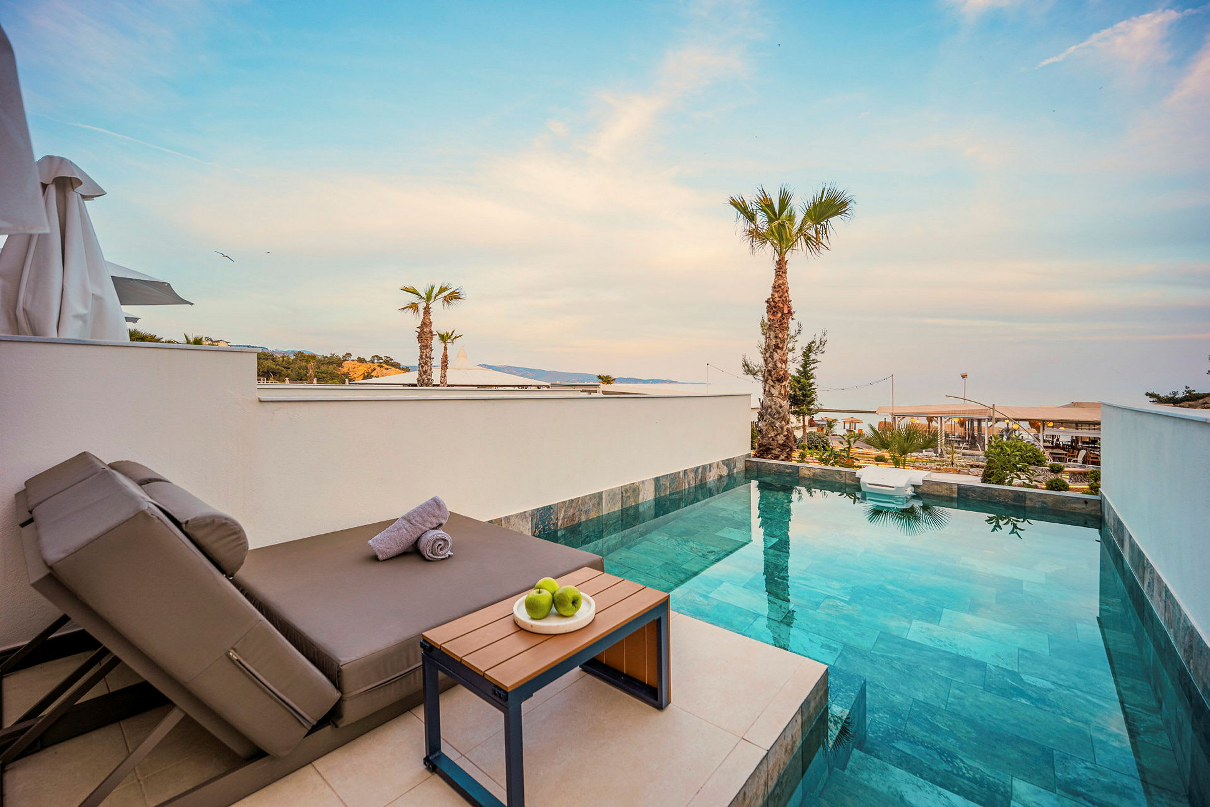 Bild av en terrass med en vilstol, ett litet bord med gröna äpplen och en privat pool med utsikt över ett palmbeklätt område. (AI-genererad text)