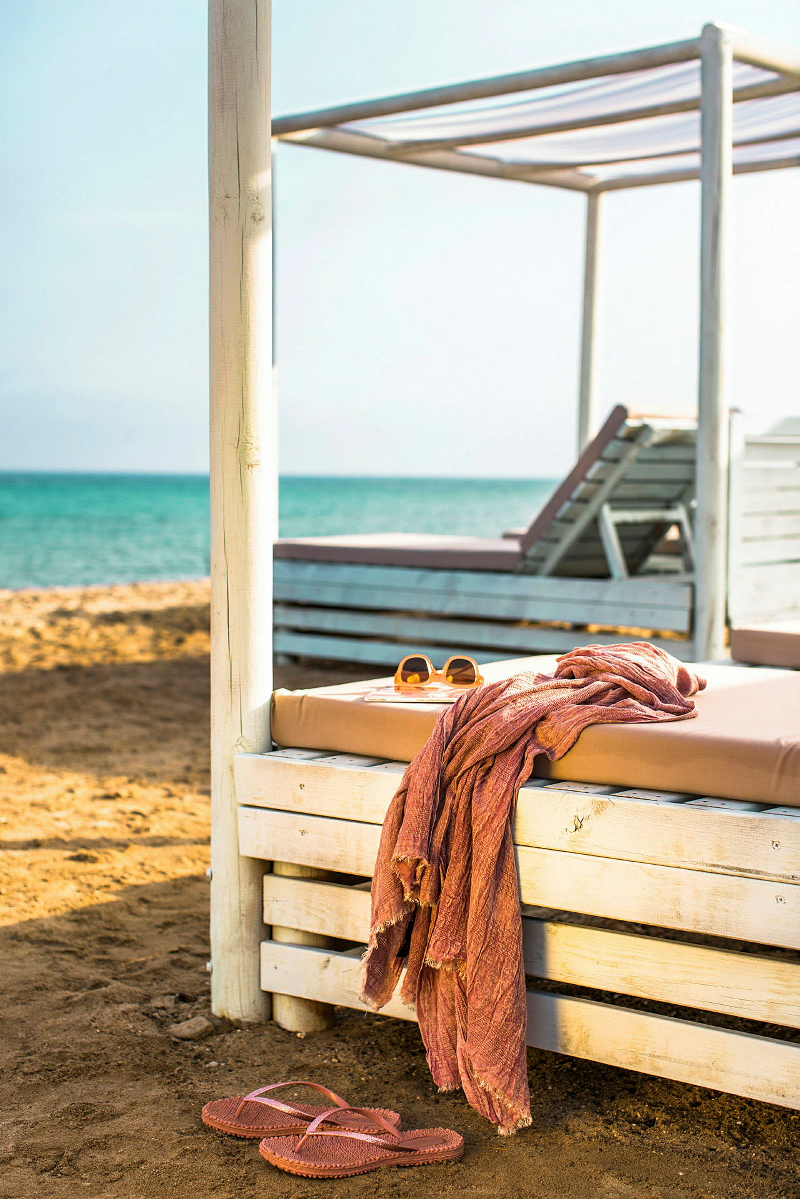 Bild av en strandbädd med solglasögon och ett klädesplagg draperat över kanten samt sandaler på marken, med havet synligt i bakgrunden. (AI-genererad text)