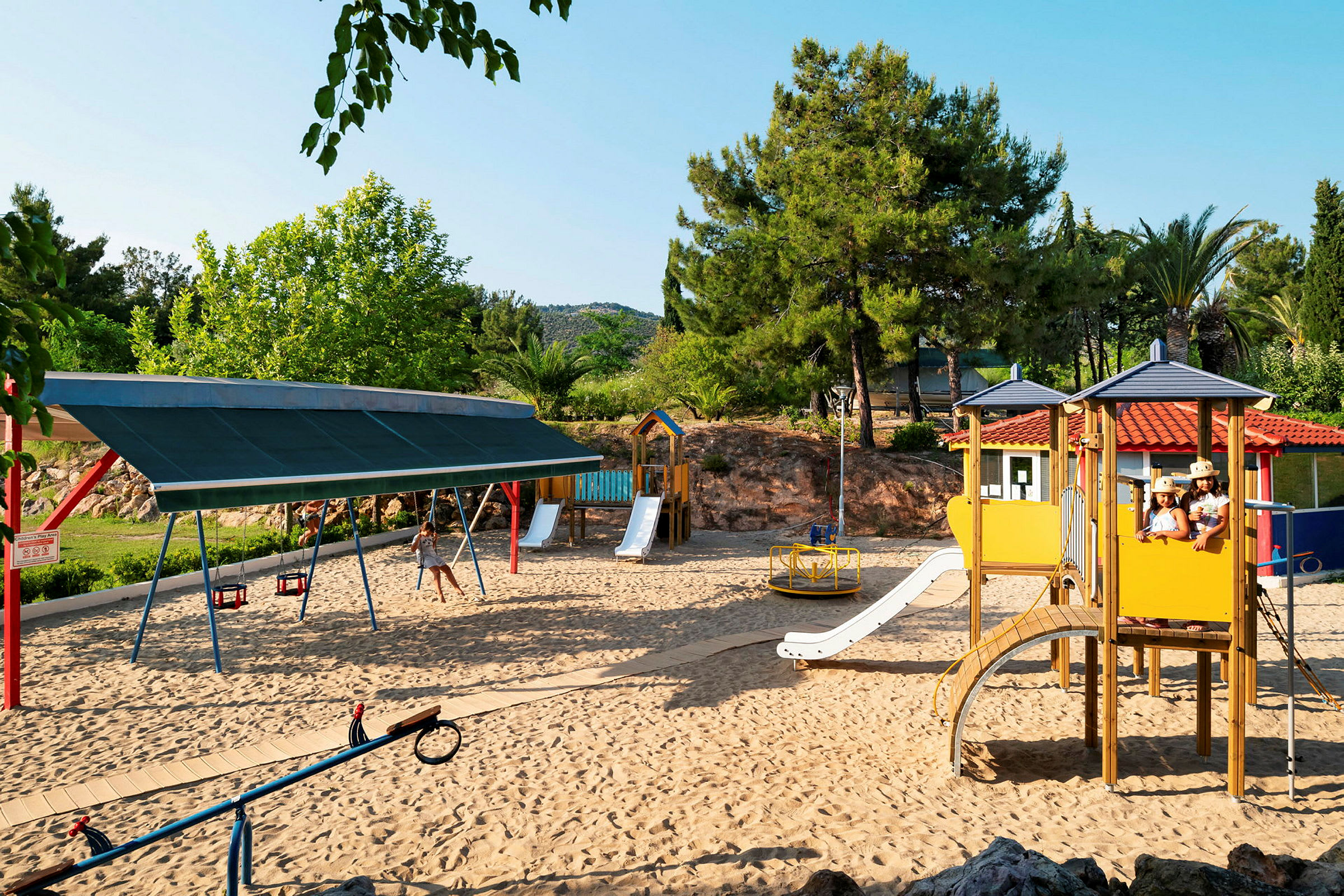 Lekplats med sand, gungor och lekställning omgiven av grönska. (AI-genererad text)