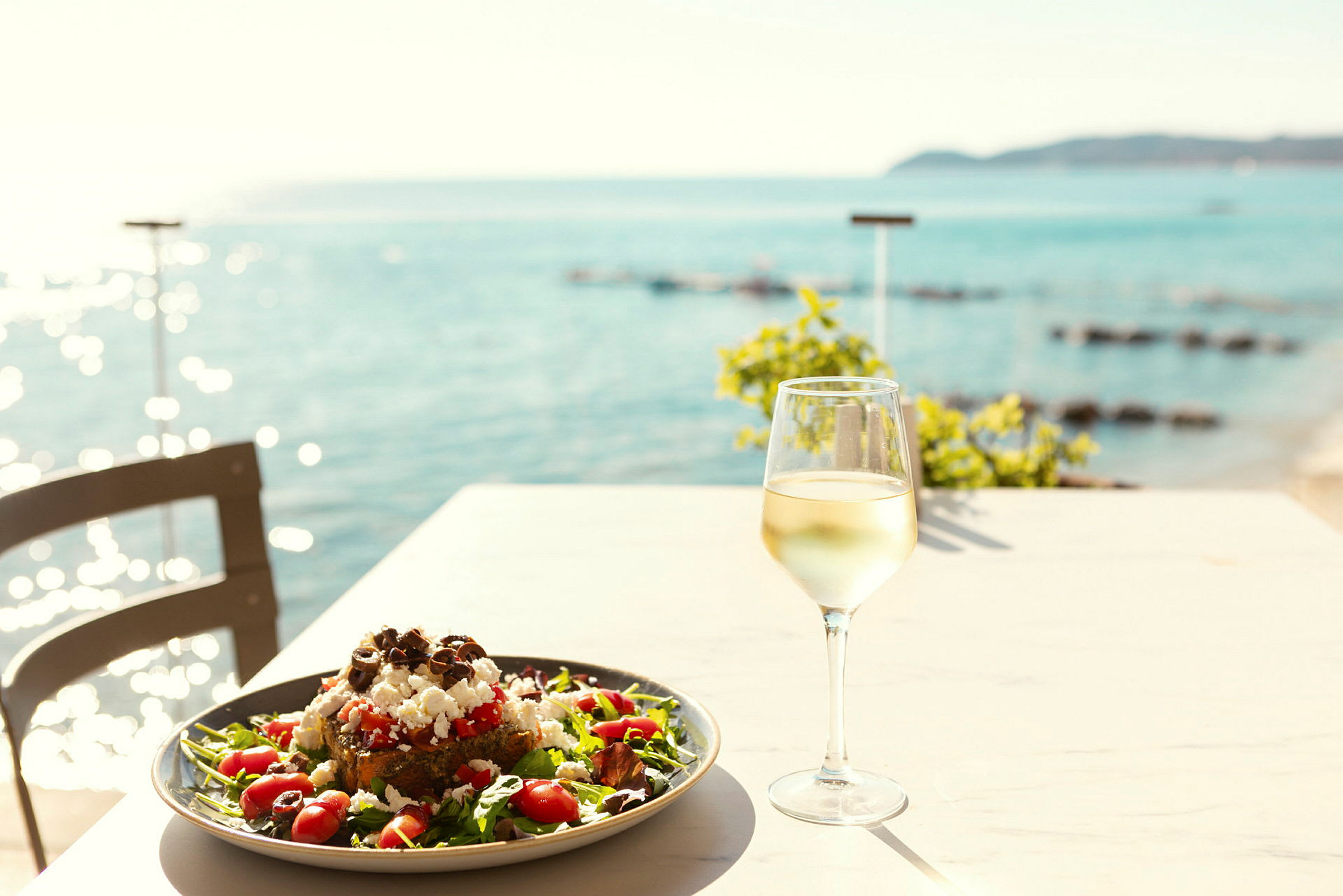 En tallrik med sallad och ett glas vitt vin på ett bord med utsikt över havet. (AI-genererad text)