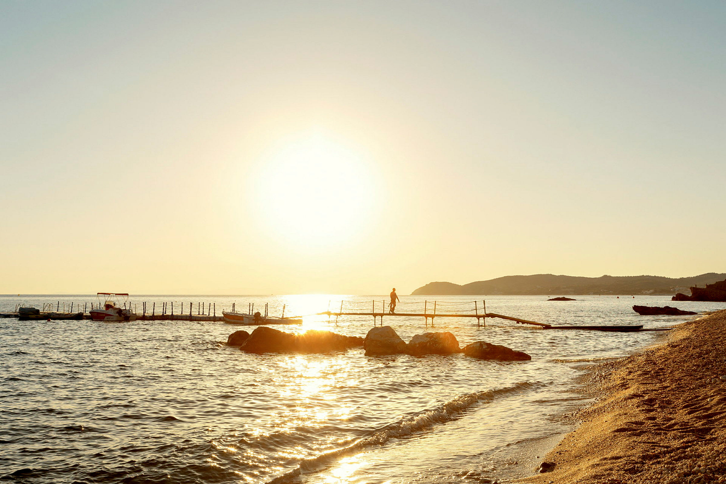 Solnedgång vid havet med brygga, stenstrand och båtar. (AI-genererad text)