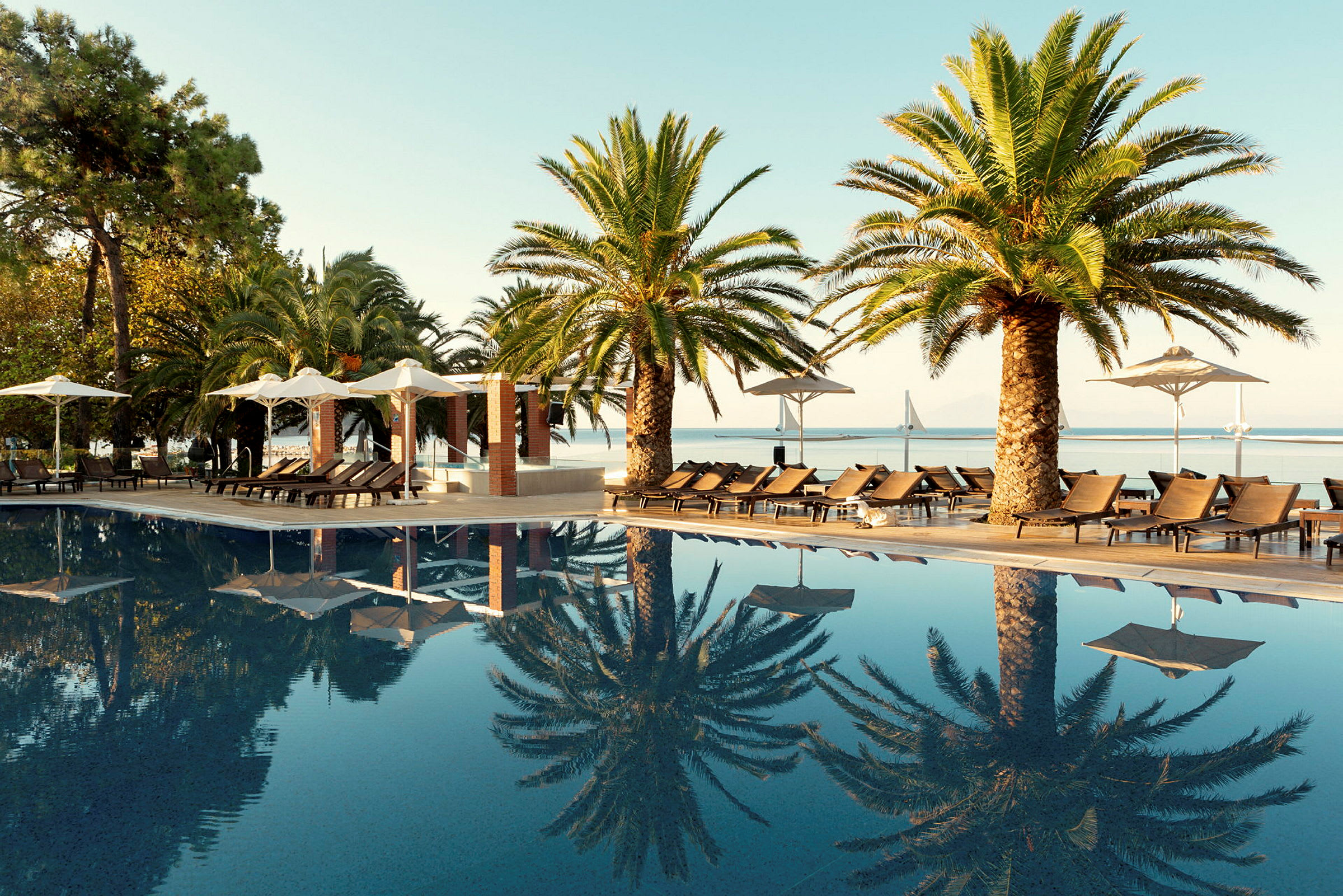 En pool med solsängar och palmer vid sidan samt utsikt mot en bro och hav. (AI-genererad text)