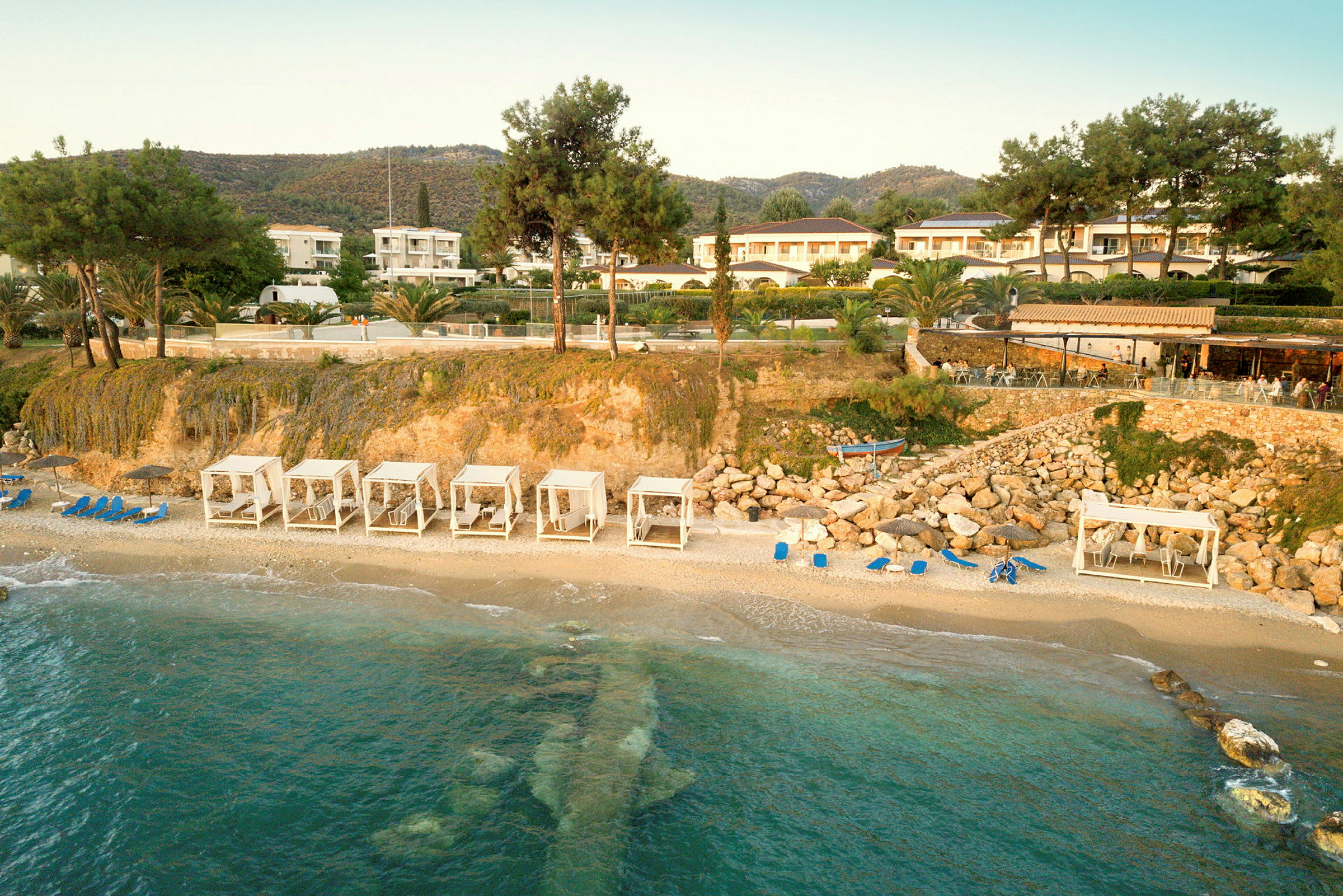 Strand med cabanas och solstolar, omgiven av gröna kullar och byggnader. (AI-genererad text)