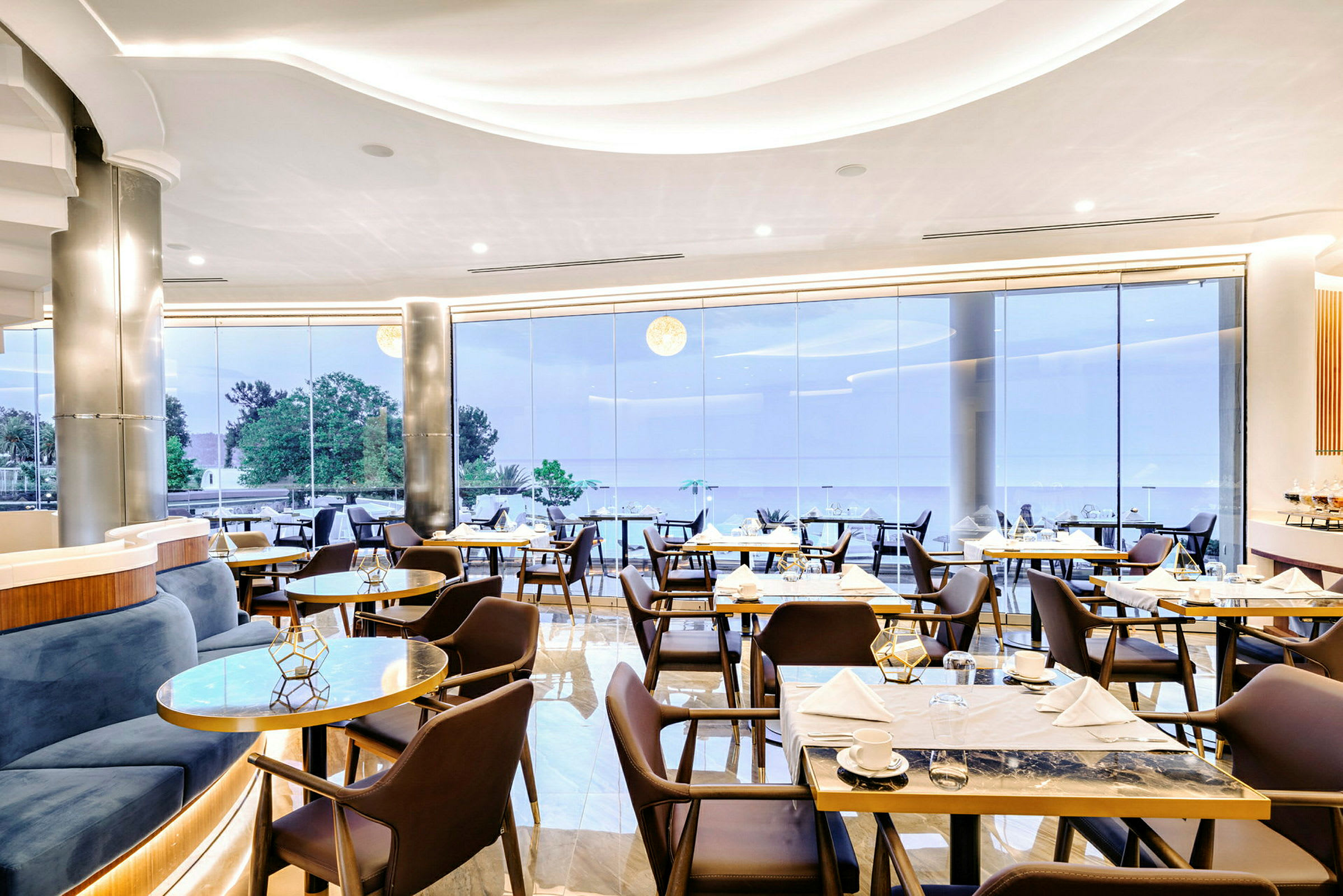 Restaurang med sittplatser, bord och stora glaspartier som visar en utsikt. (AI-genererad text)