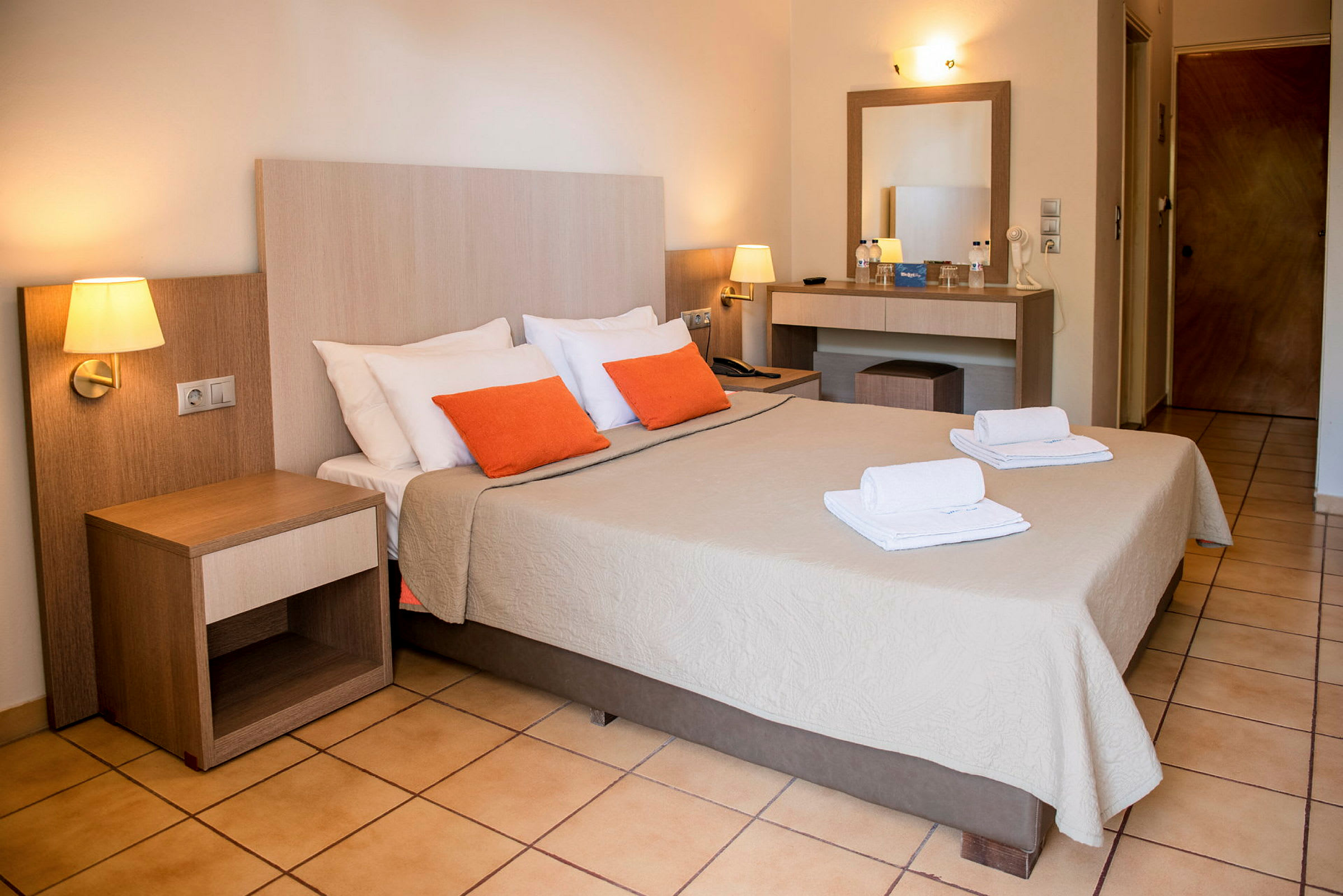 Hotellrum med dubbelsäng, två orange kuddar och vita handdukar på sängen. (AI-genererad text)