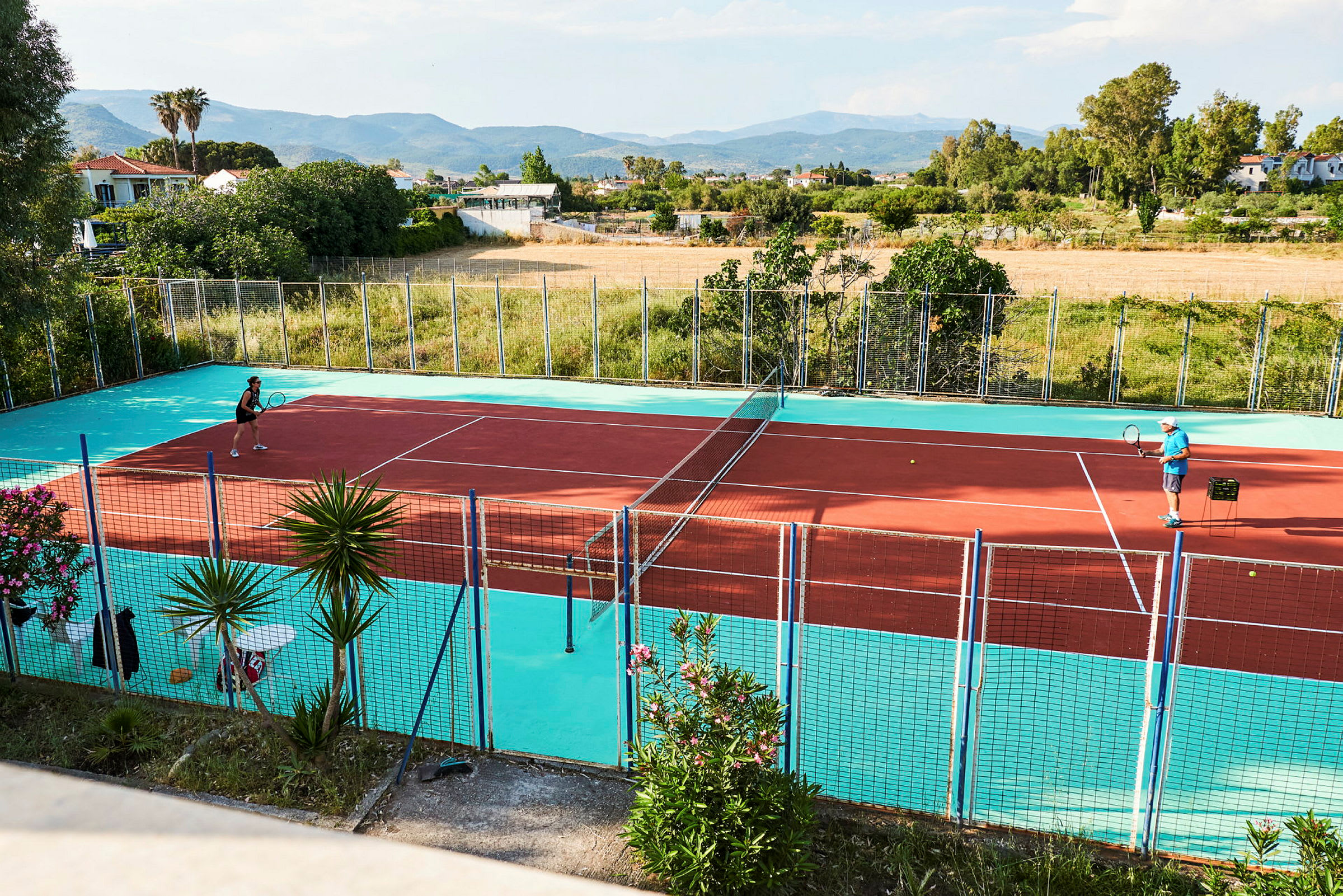 Två personer spelar tennis på en färgglad tennisbana utomhus med landskap i bakgrunden. (AI-genererad text)