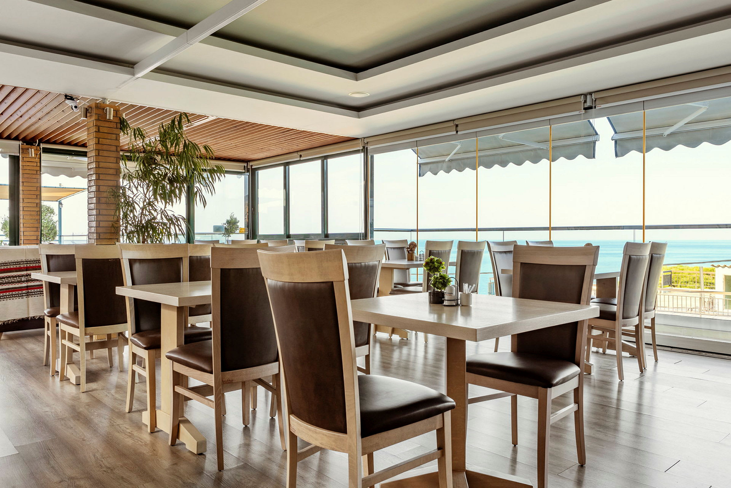Interiör av en restaurang med träbord och stolar, stora fönster med utsikt över havet. (AI-genererad text)