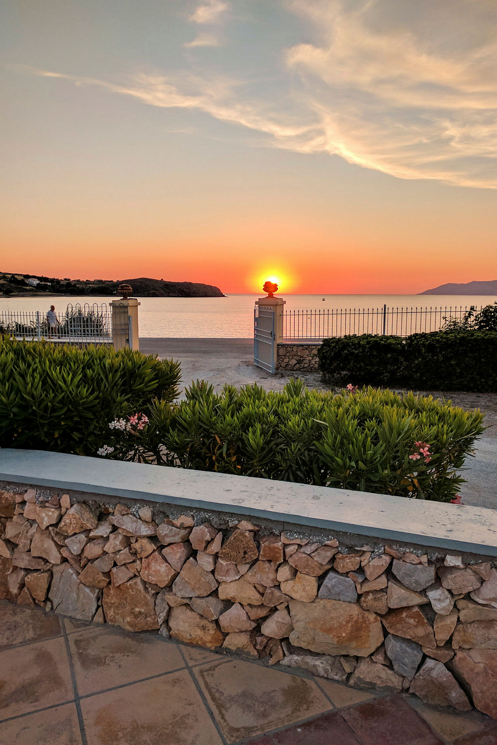 Solnedgång sett över havet med en stengärdsgård och grönska i förgrunden. (AI-genererad text)