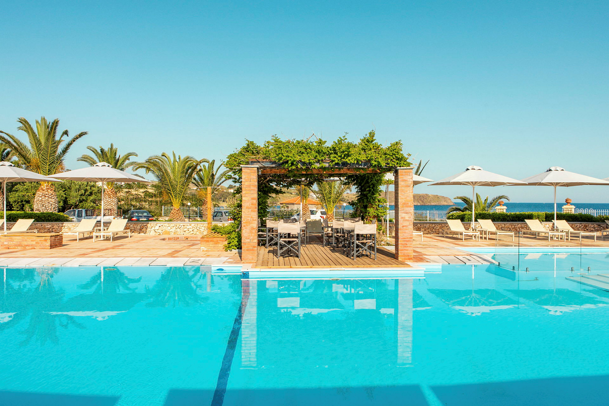 En swimmingpool omgivet av solstolar, parasoller och en pergola vid kusten. (AI-genererad text)