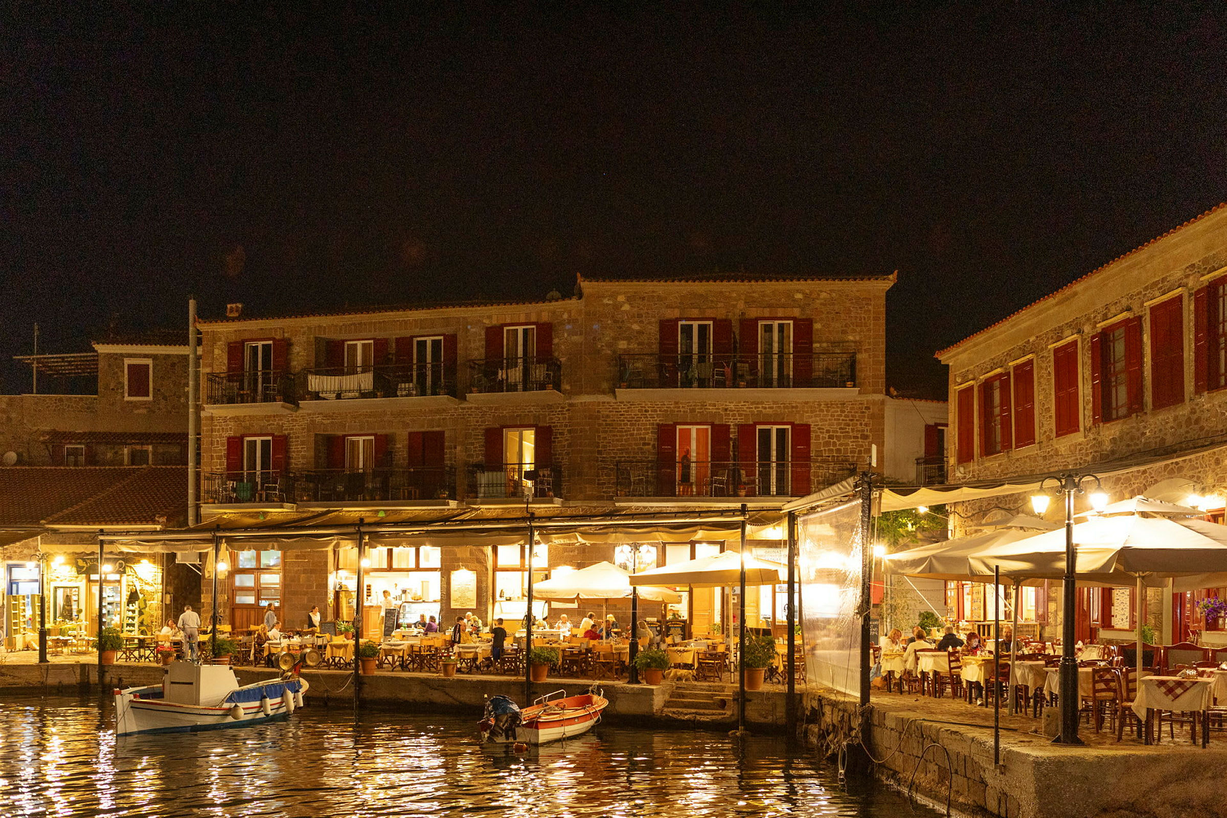 En kvällsvy av en hamn med byggnader och restaurangområden upplysta, båtar ligger vid kajen. (AI-genererad text)