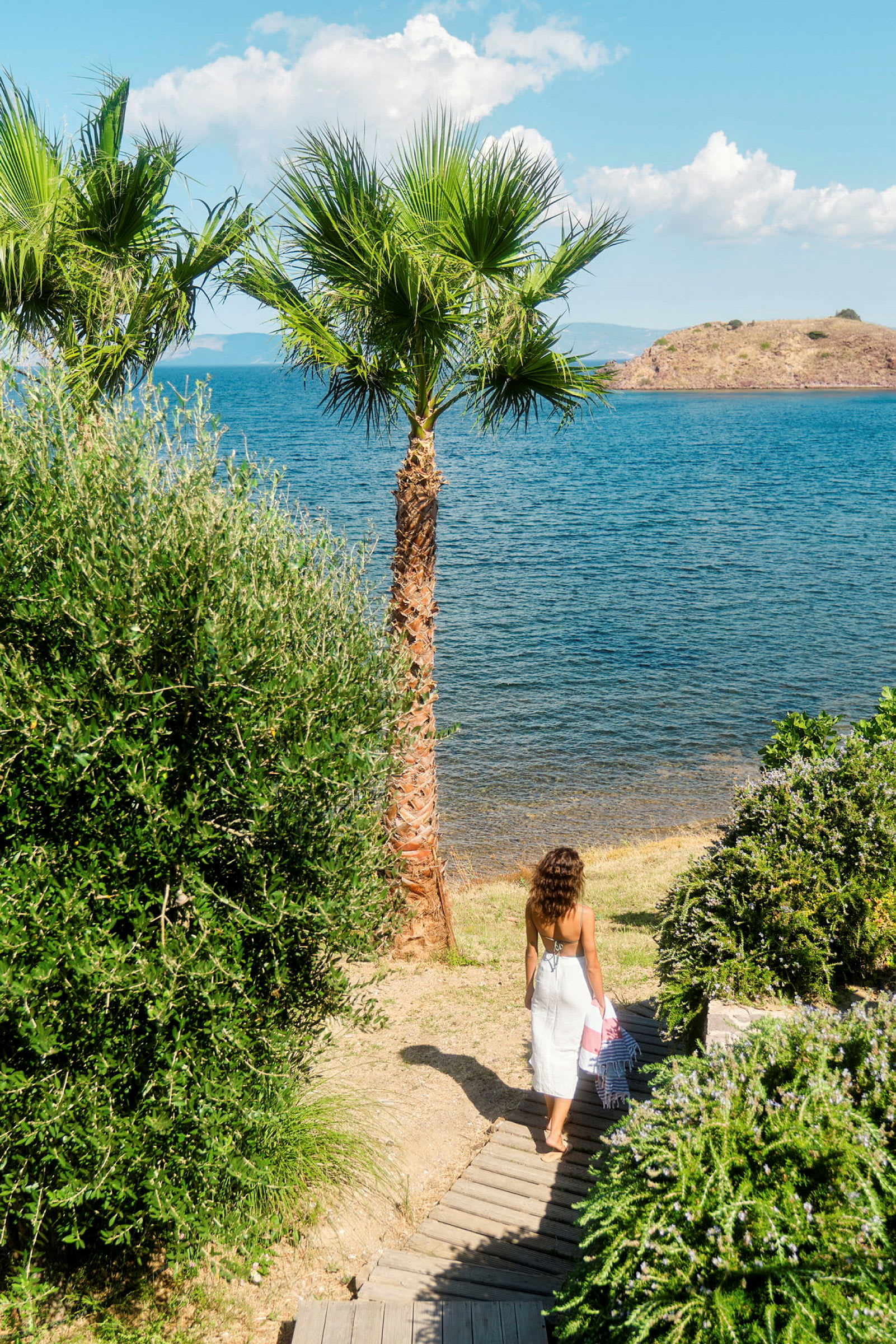 En kvinna går ner mot en strand på en trästig, omgiven av grönska och palmer. (AI-genererad text)