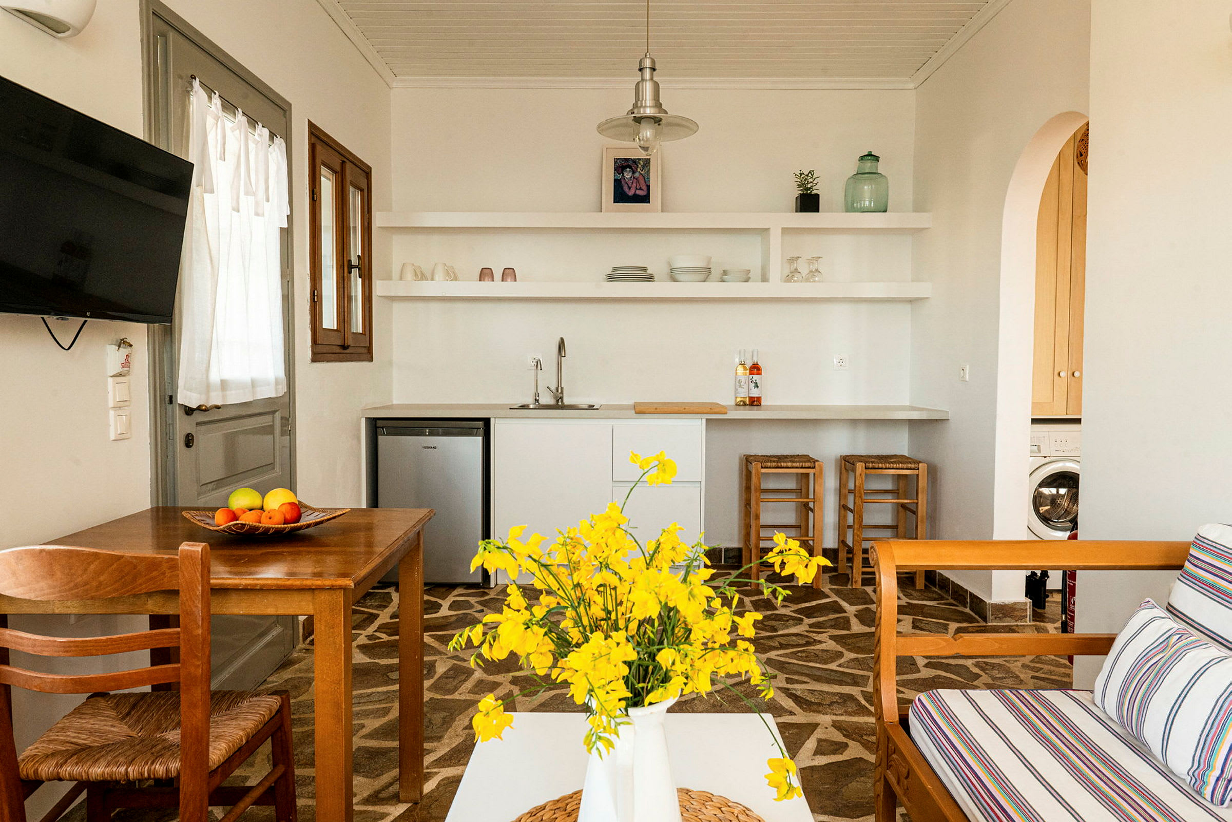 Ett ljust inrett kök med vita hyllor, ett matbord i trä, gula blommor i en vit vas och ett vardagsrum med träsoffa och randiga dynor. (AI-genererad text)