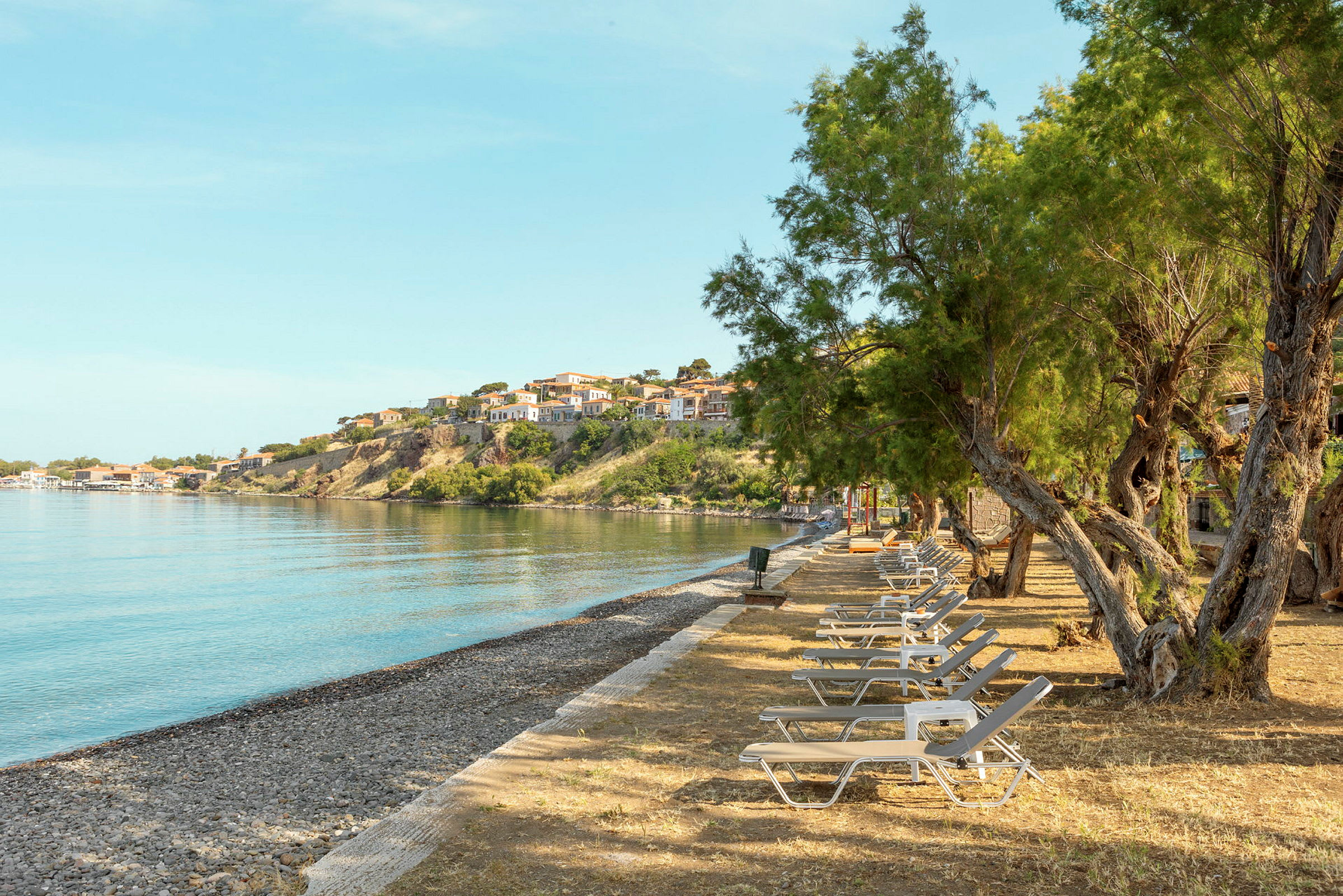 En strand med solstolar och träd i förgrunden, med hus på en sluttning vid vattnet i bakgrunden. (AI-genererad text)