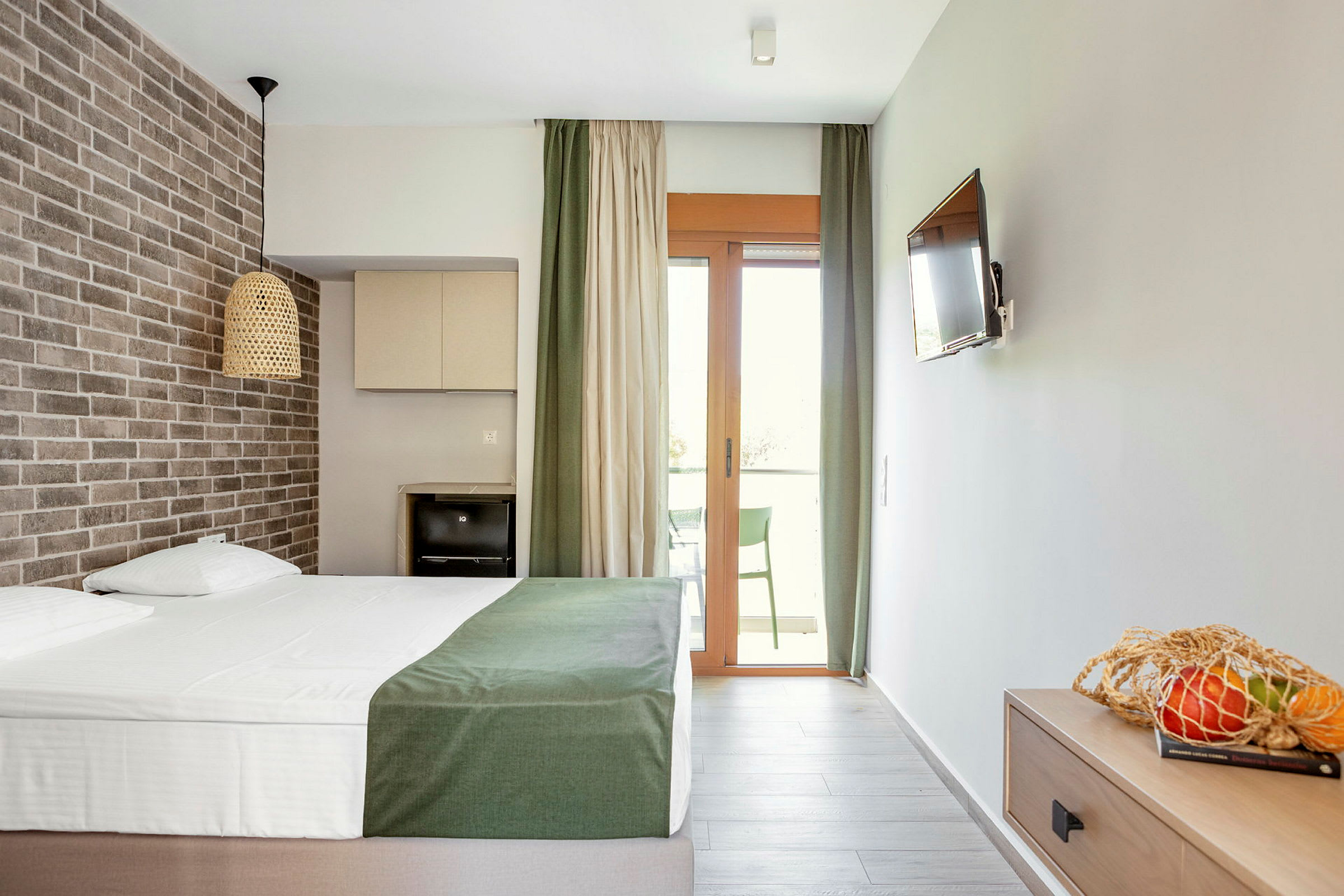 Ett hotellrum med en dubbelsäng, ett tegelvägg, en TV, en balkong och en korg med frukt på ett bord. (AI-genererad text)