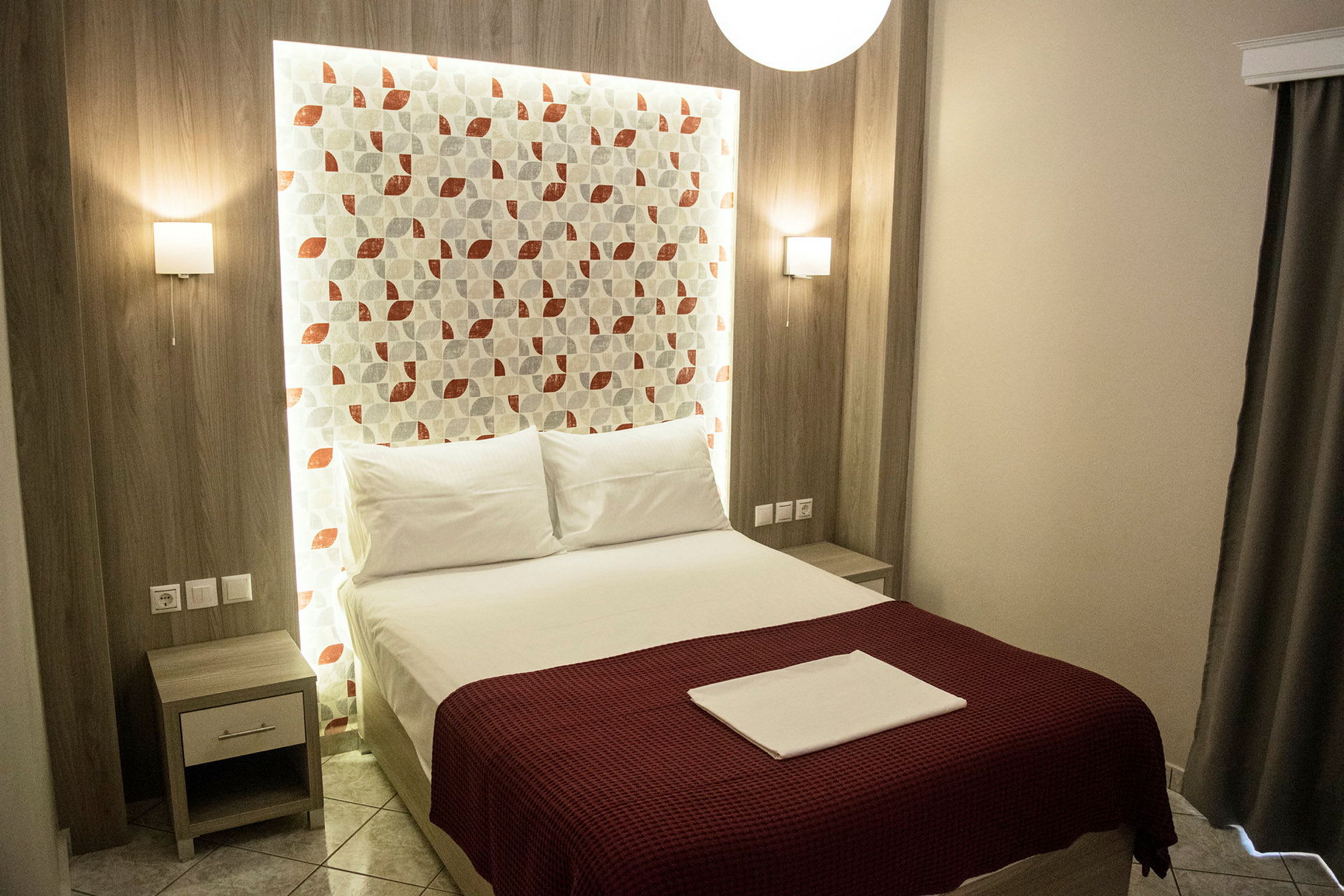 En modern hotellrum med en säng som har röda sängkläder och ett huvudgavel med geometriskt mönster. (AI-genererad text)