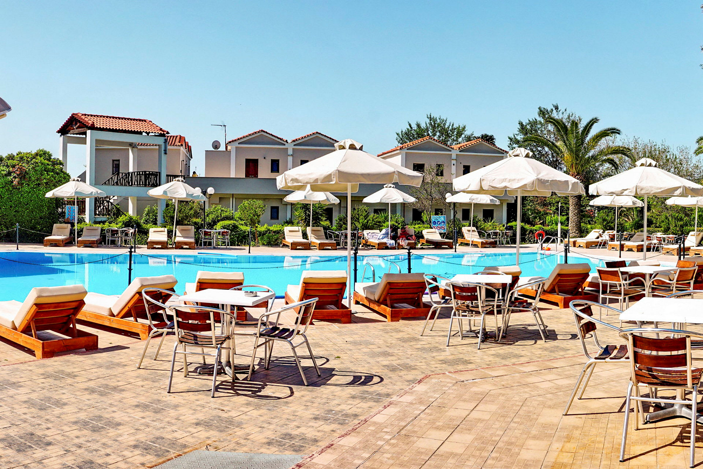 En hotellpool omgiven av solstolar, parasoller och byggnader. (AI-genererad text)