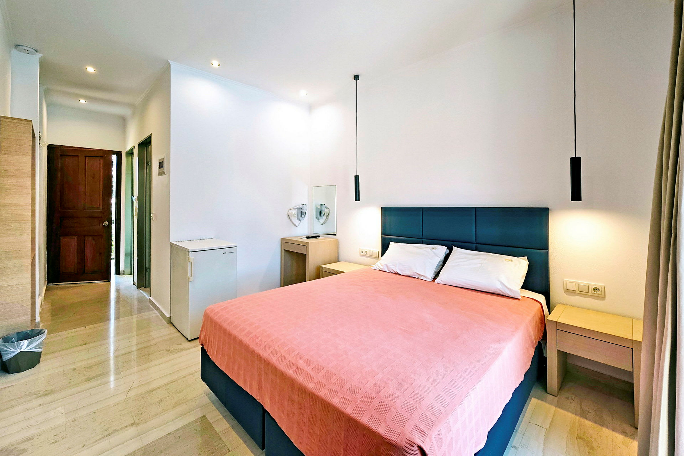 Ett hotellrum med en dubbelsäng och rosa överkast, omgiven av möbler och ett fönster. (AI-genererad text)