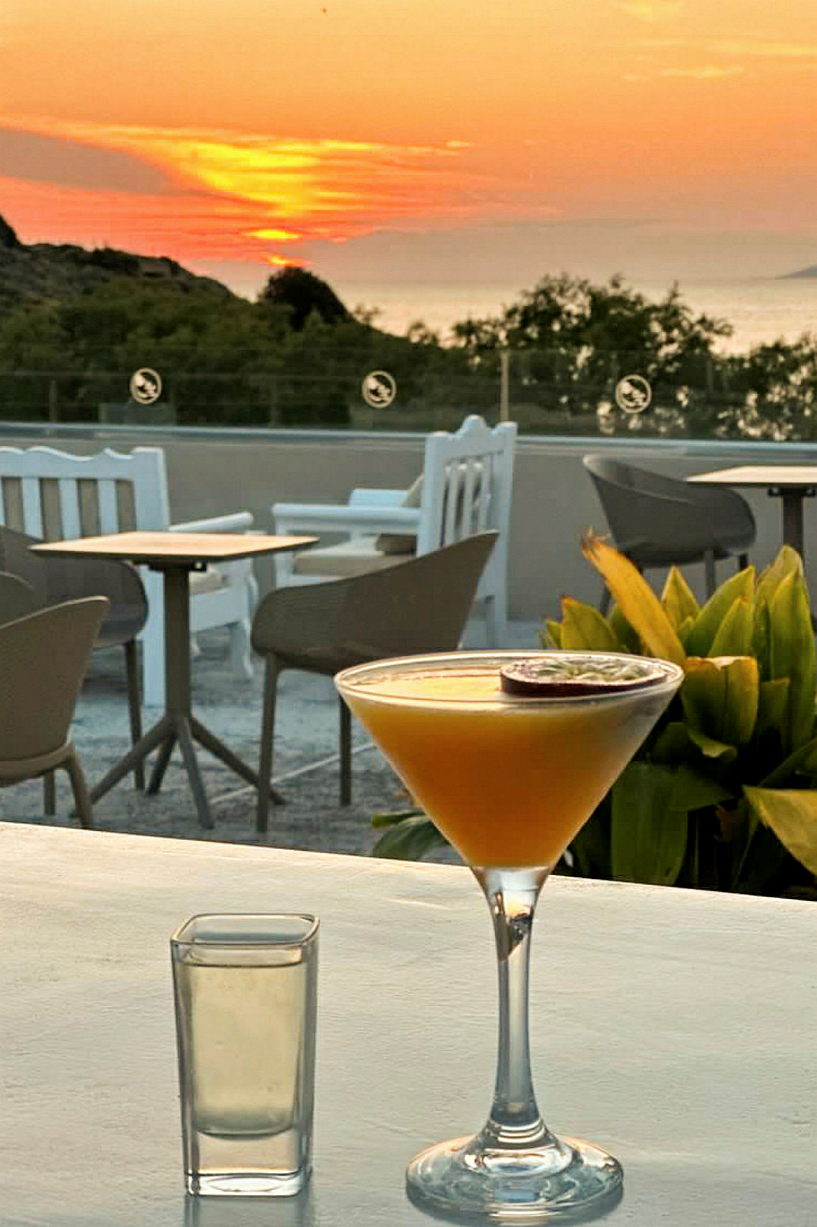 En tropisk cocktail och ett glas står på ett bord vid solnedgång med sittmöbler och grönska i bakgrunden. (AI-genererad text)
