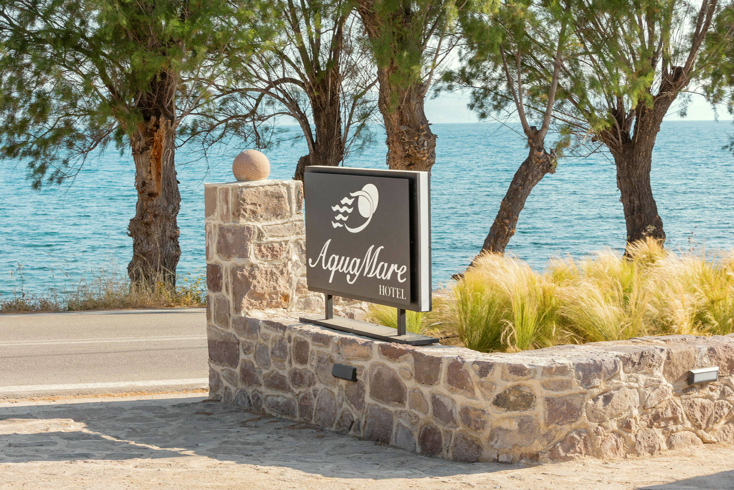 Hotellskylt med texten 'Aqua Mare Hotel', omgiven av stenmur och växter, med havet och träd i bakgrunden. (AI-genererad text)