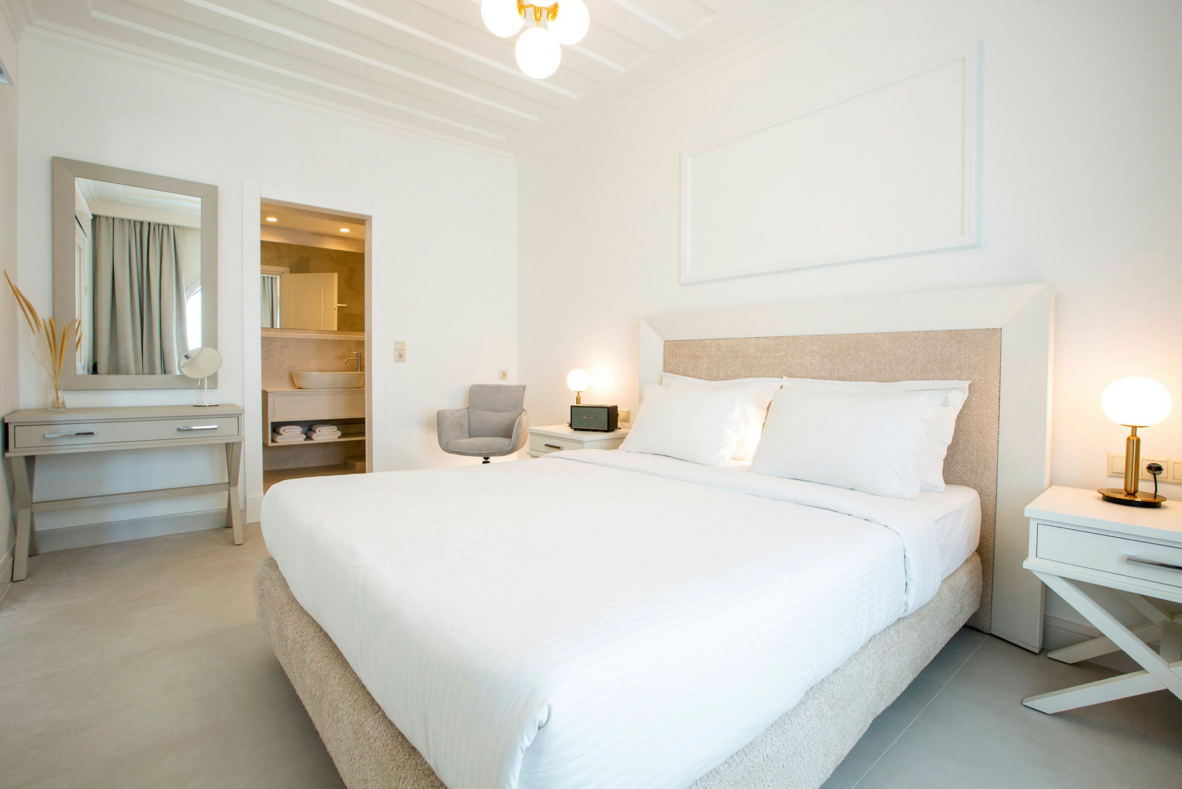 Ett ljust och modernt hotellrum med en stor säng, två nattduksbord med lampor, ett skrivbord med spegel, och ett öppet badrum. (AI-genererad text)