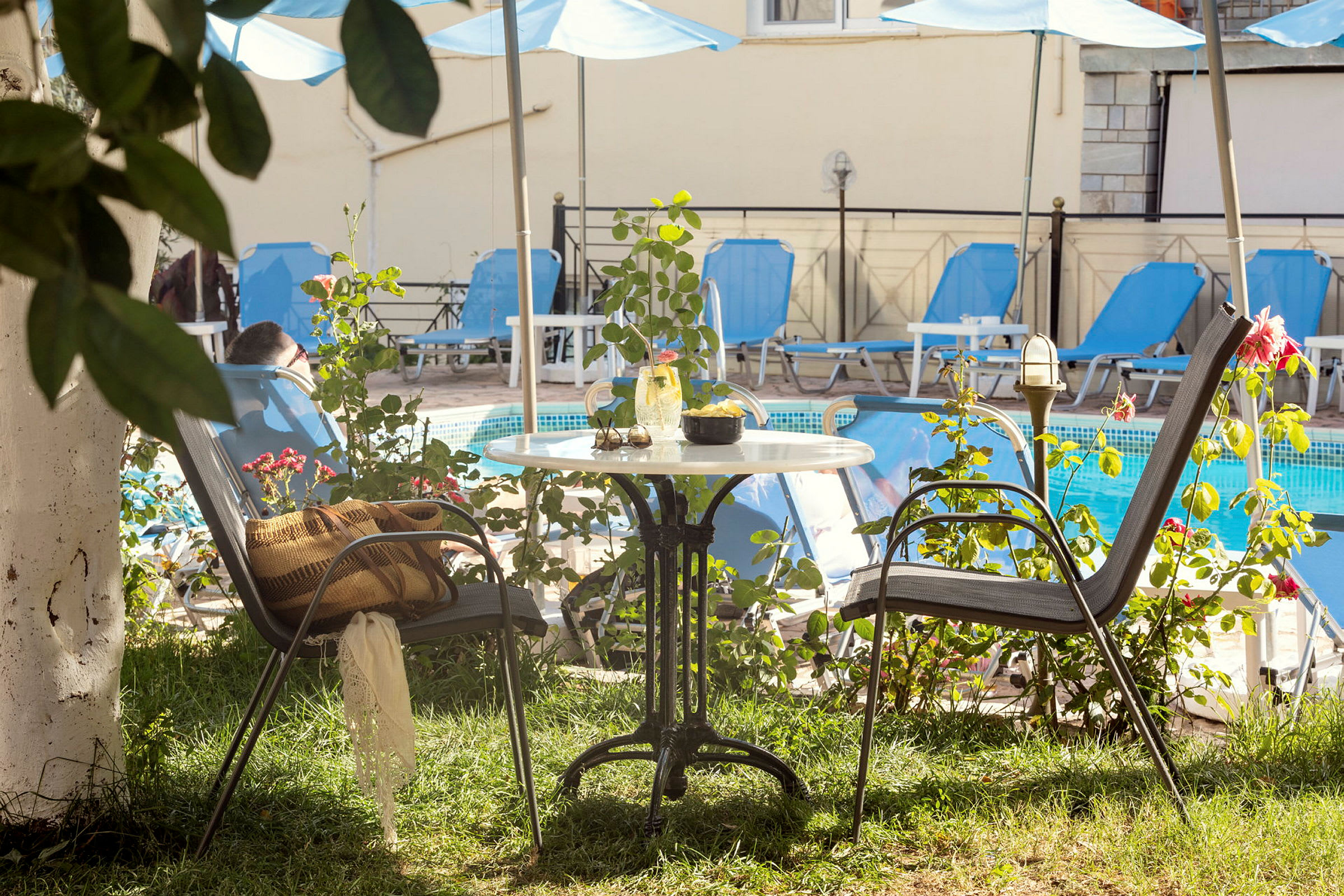 Ett bord med två stolar står i en trädgård bredvid en pool och solstolar under parasoll. (AI-genererad text)