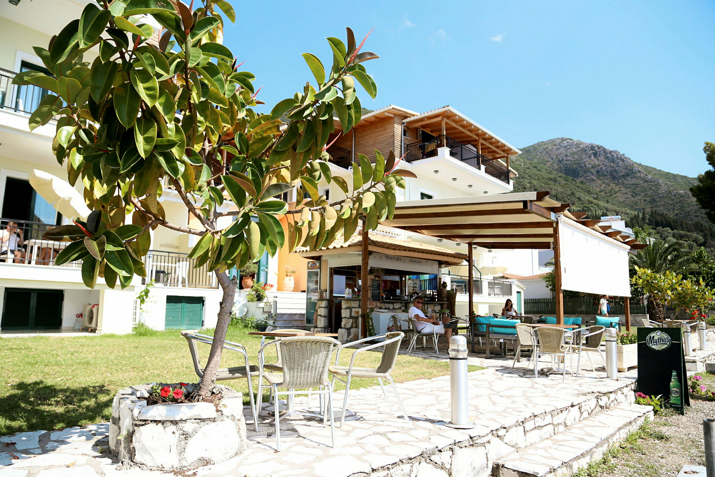 En utomhusrestaurang framför ett mindre lägenhetshotell med en bergssluttning i bakgrunden. (AI-genererad text)