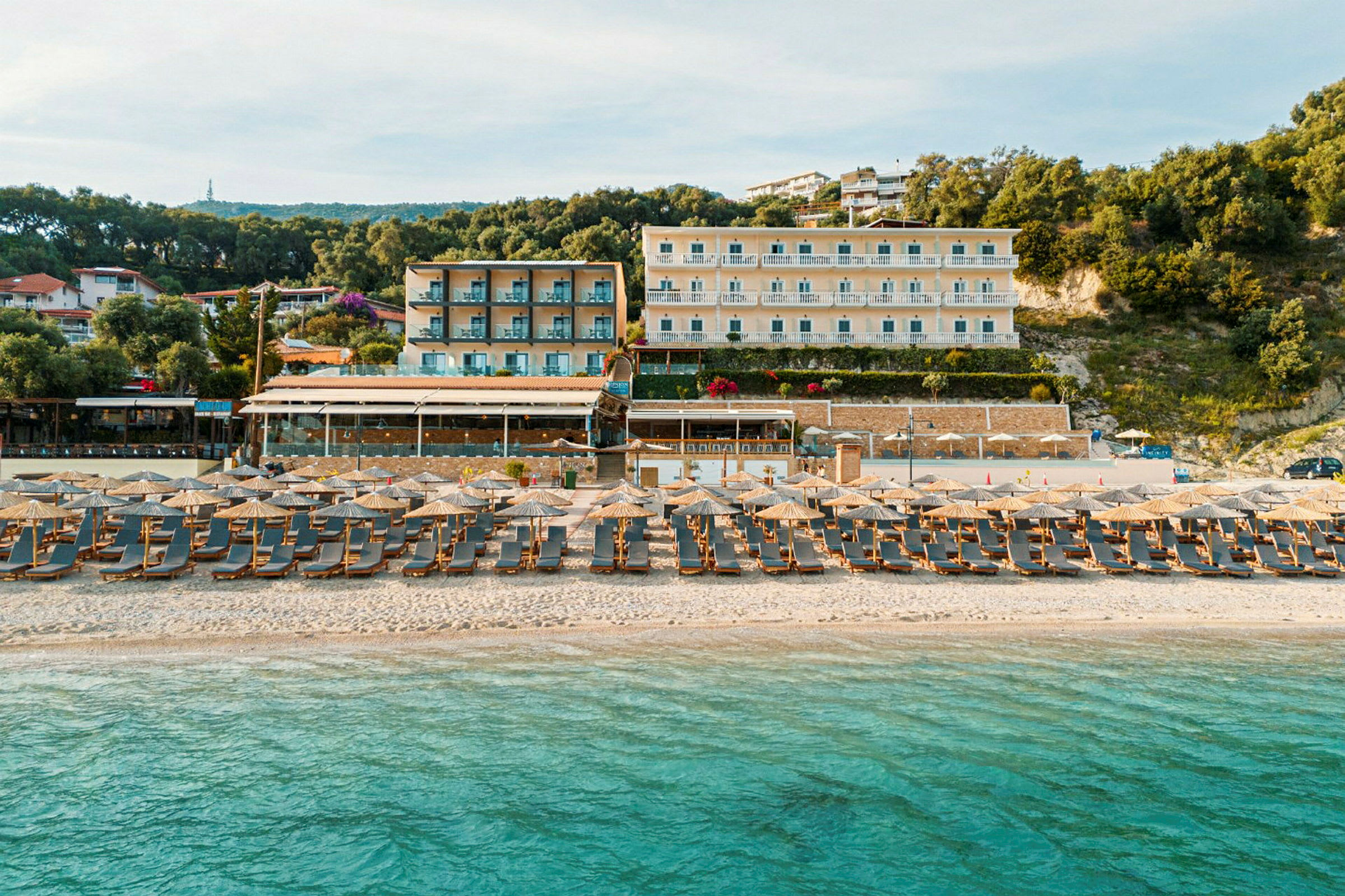 En sandstrand vid havet med solstolar och parasoller. Bakgrunden visar hotellbyggnader och grönska. (AI-genererad text)