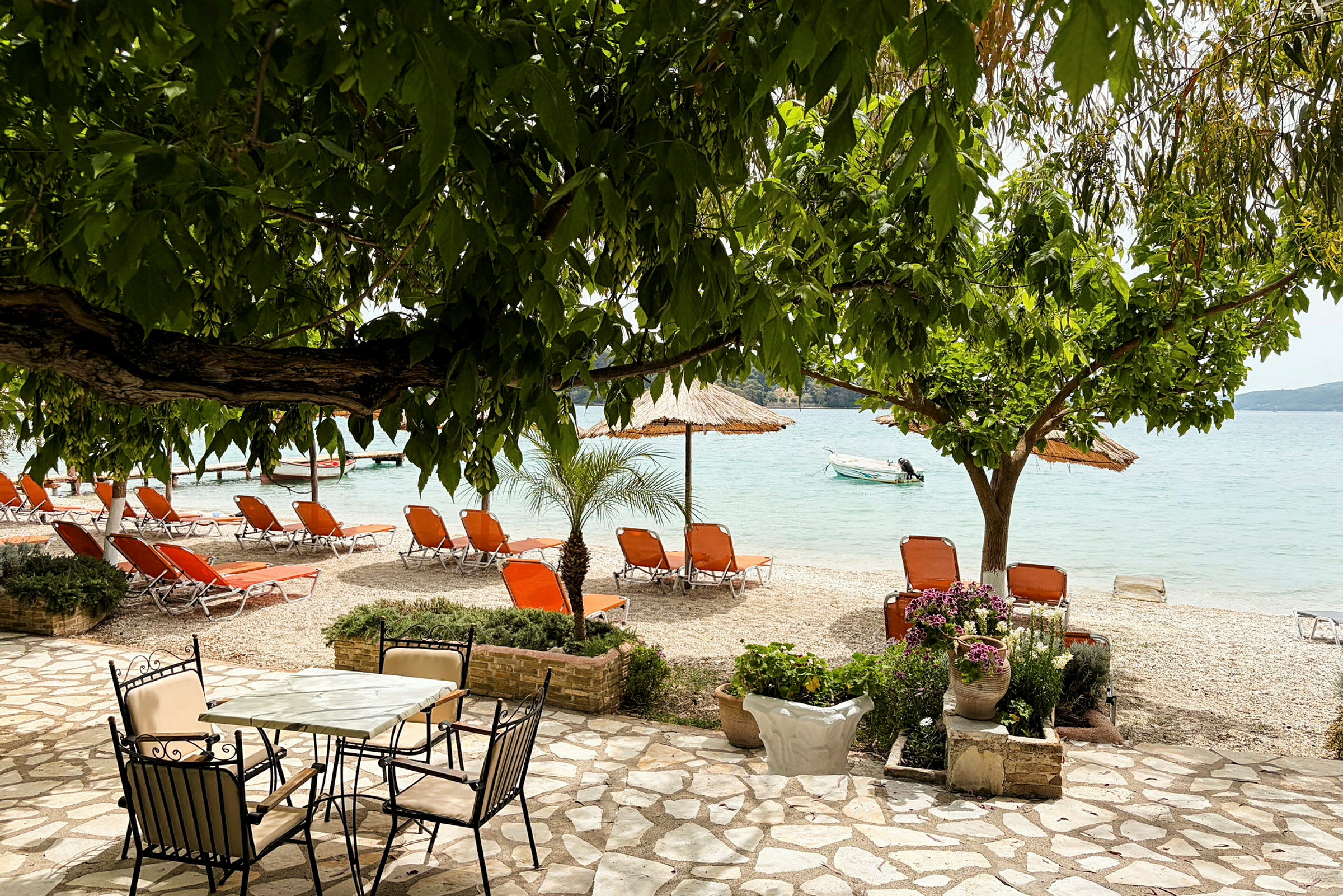 En strand med orange solstolar, parasoller och vy ut mot havet. En stenlagd yta med bord och stolar samt planterade blommor i förgrunden. (AI-genererad text)