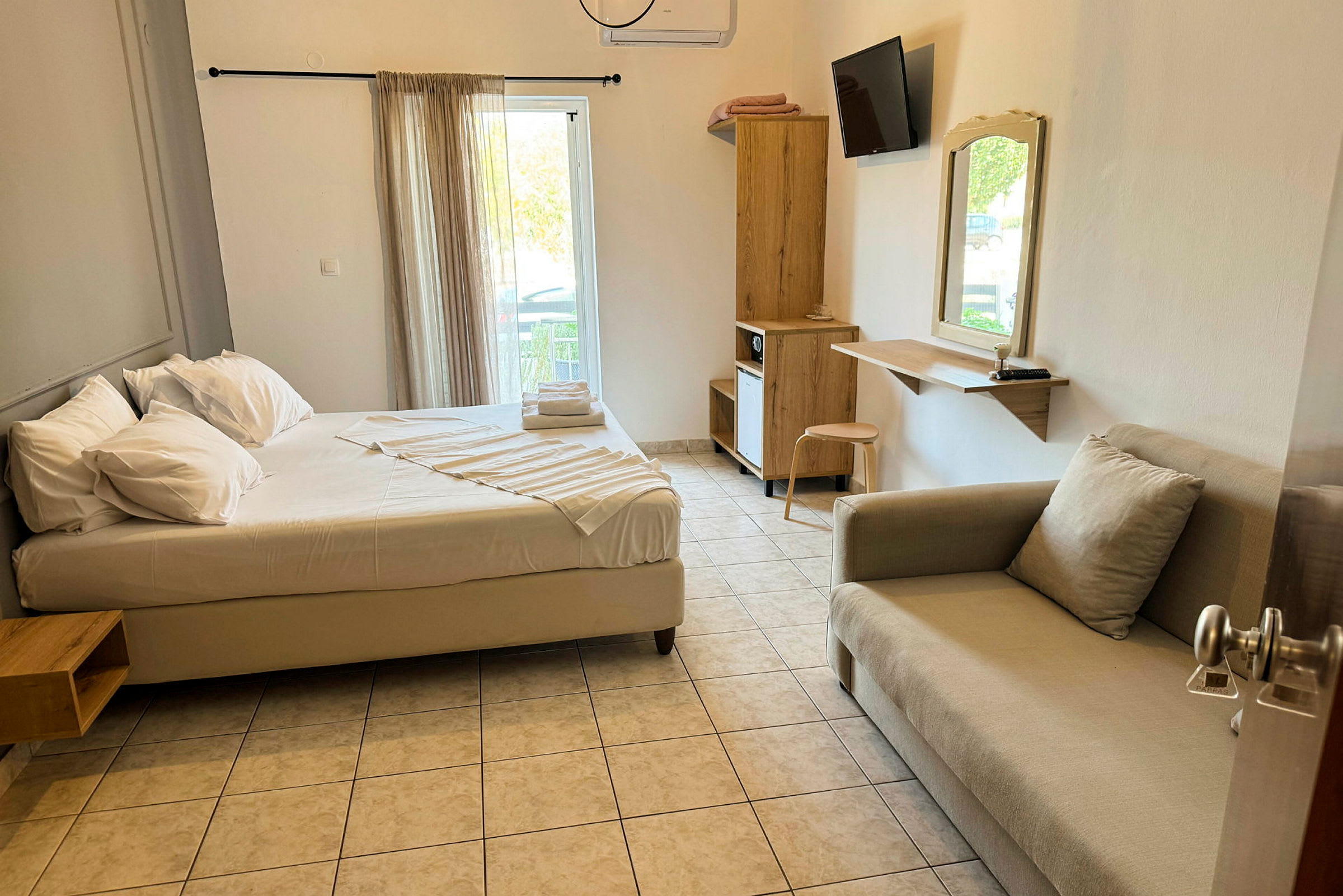 Ett hotellrum med en säng, soffa, skrivbord och garderob. Fönstret har ljusa gardiner. (AI-genererad text)