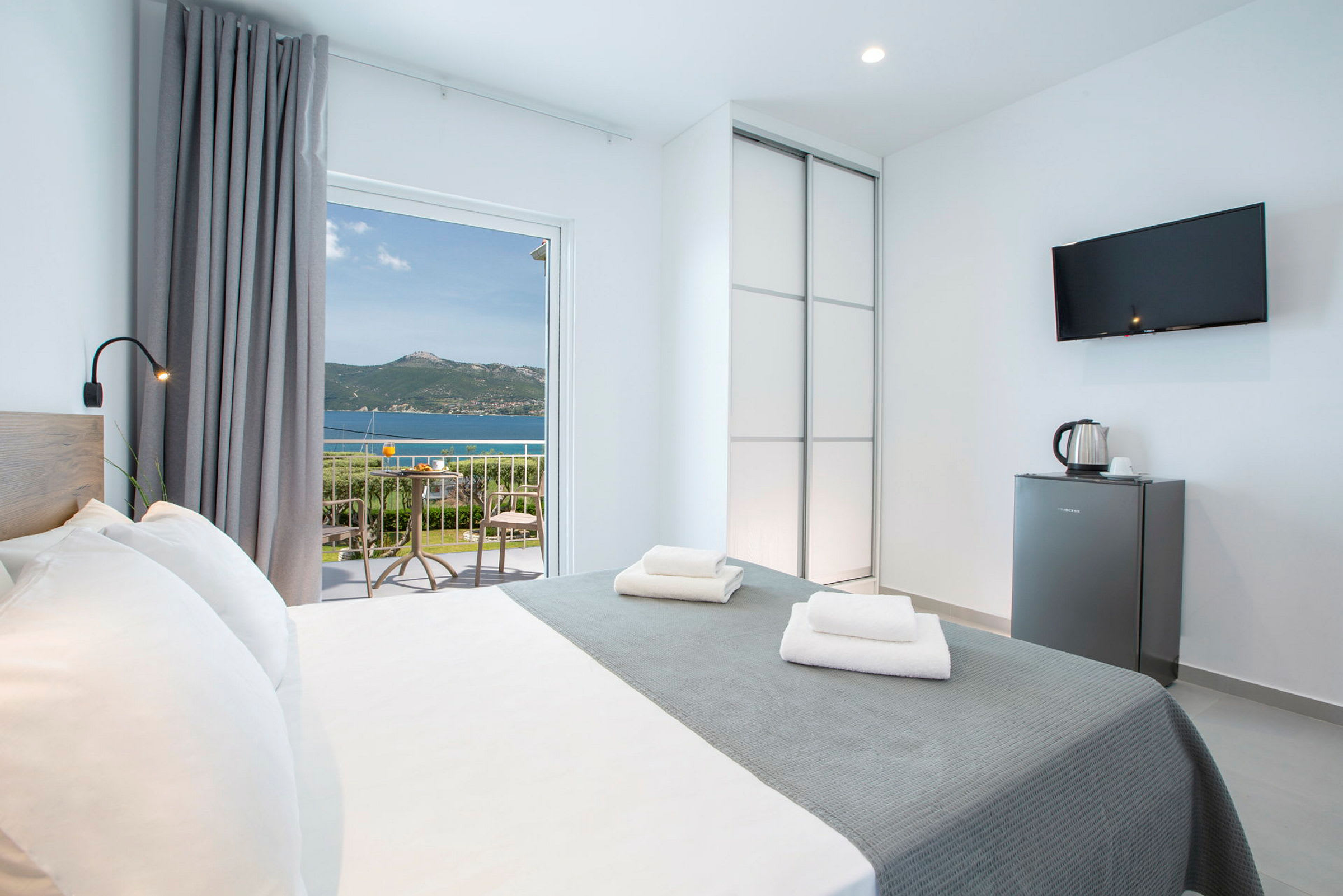 Ett hotellrum med en dubbelsäng, en garderob och en TV. Balkongen har utsikt över havet och bergen. (AI-genererad text)