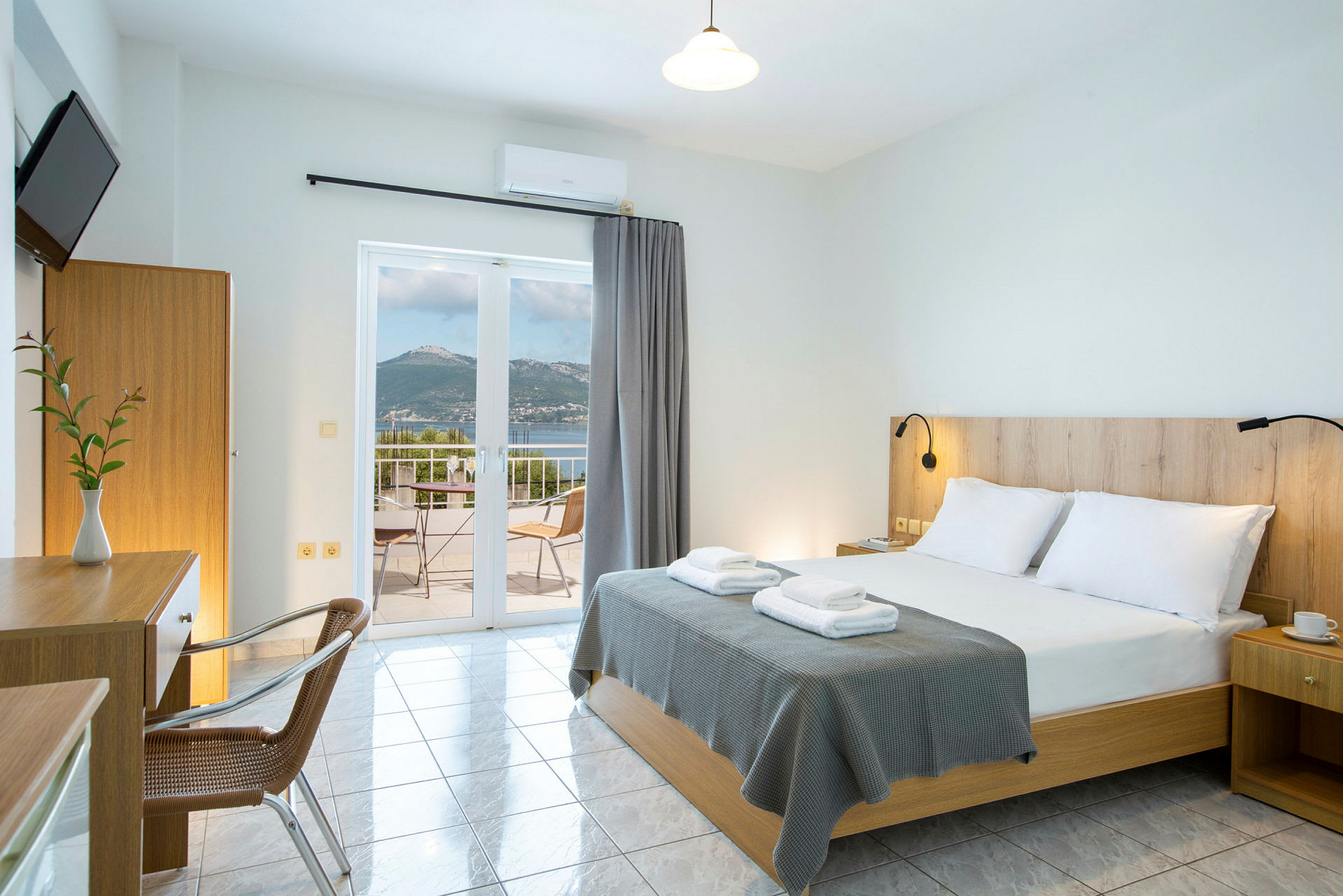 Ett hotellrum med en dubbelsäng, skrivbord och utsikt över en terrass med hav och berg. (AI-genererad text)
