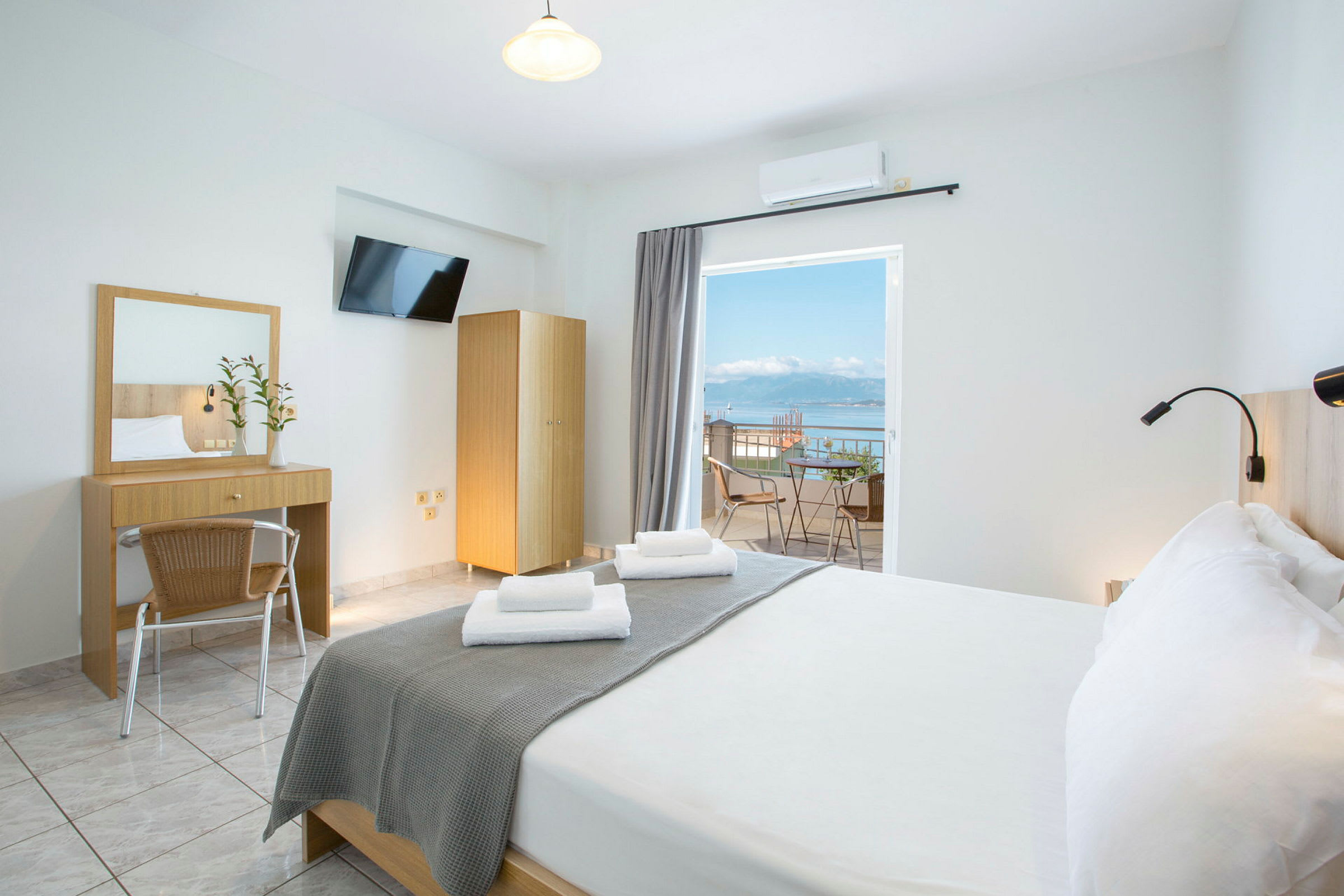 Ett ljust hotellrum med en dubbelsäng, skrivbord, spegel och en balkong med utsikt över havet. (AI-genererad text)