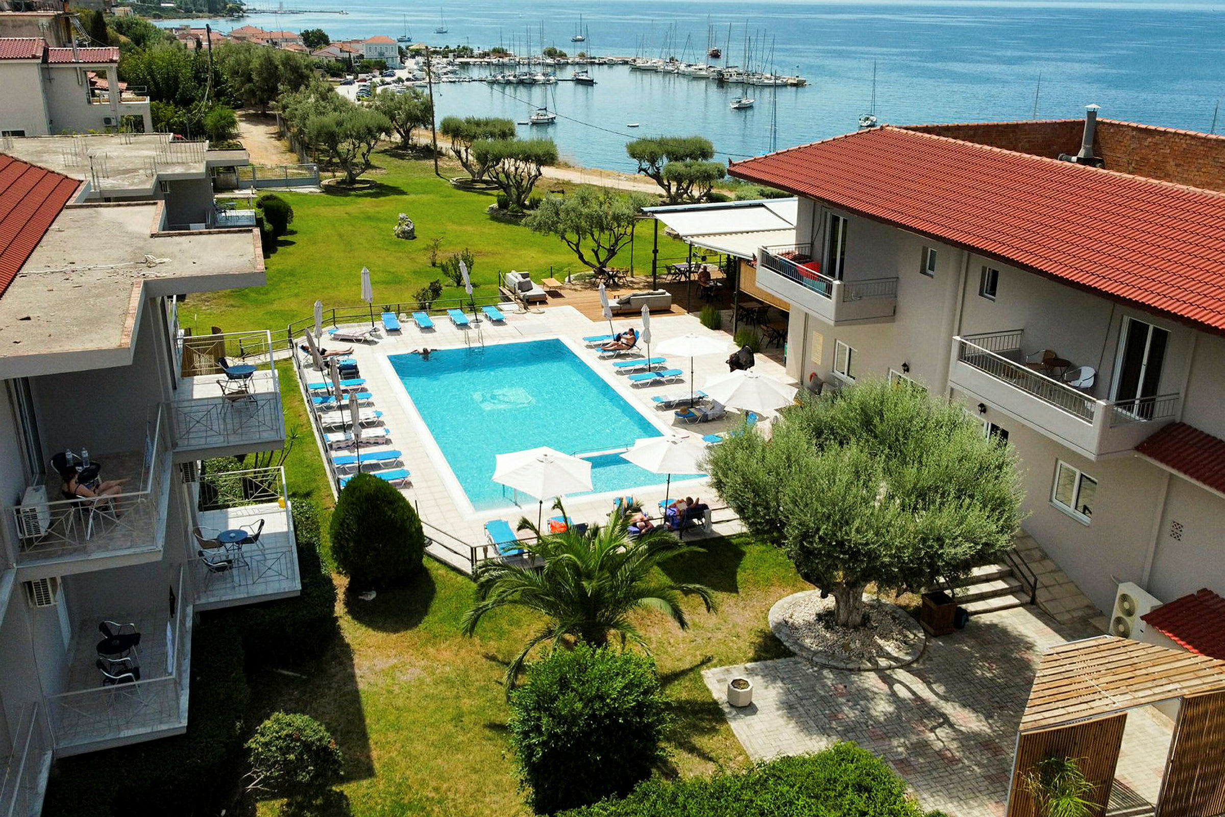 Bild på ett hotellområde med en utomhuspool, solstolar och parasoller, samt omgivande grönytor och utsikt mot en hamn vid havet. (AI-genererad text)