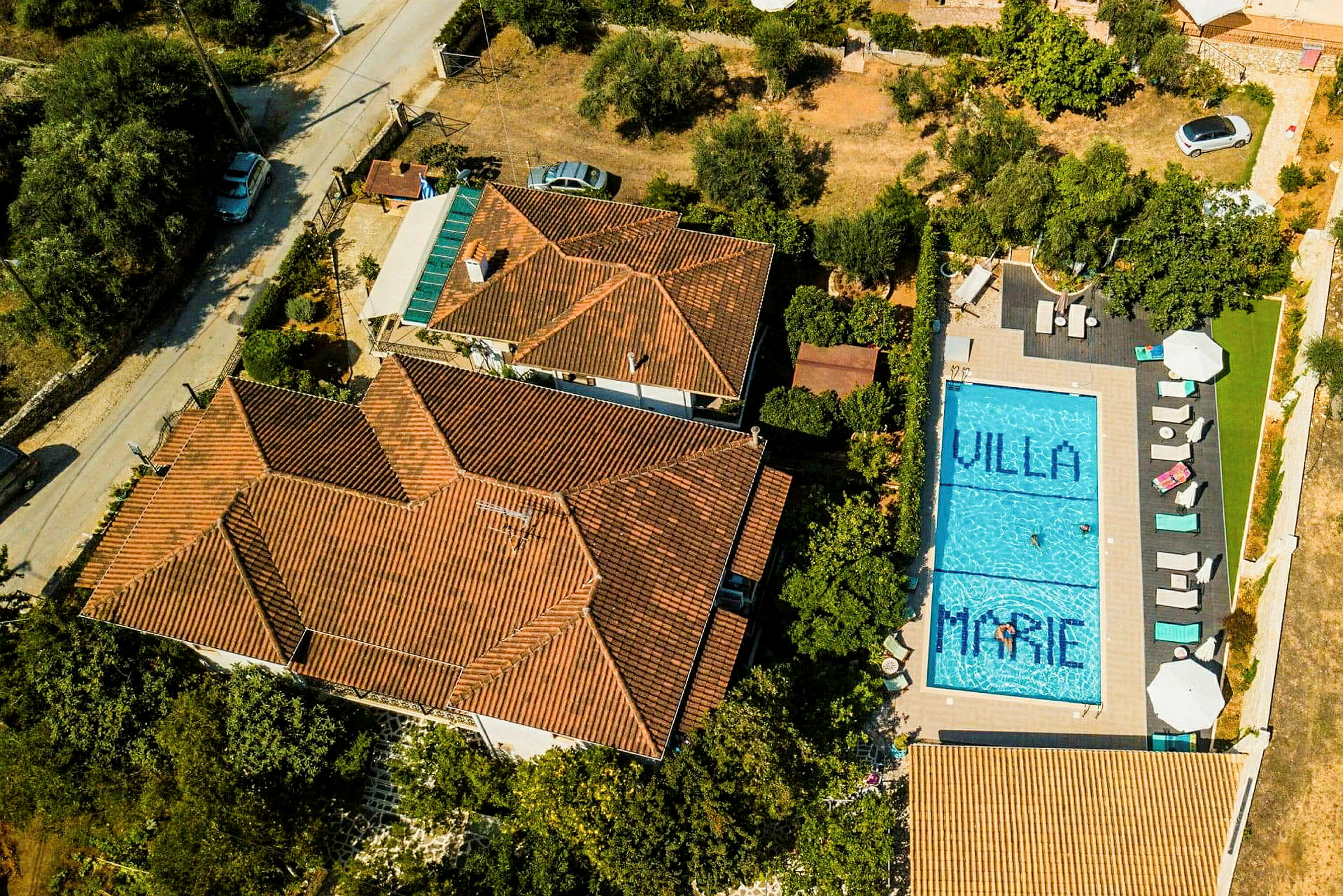 Flygfoto av en villa med orange tegeltak och en blå pool på tomten. Poolen har texten 'Villa Marie'. (AI-genererad text)