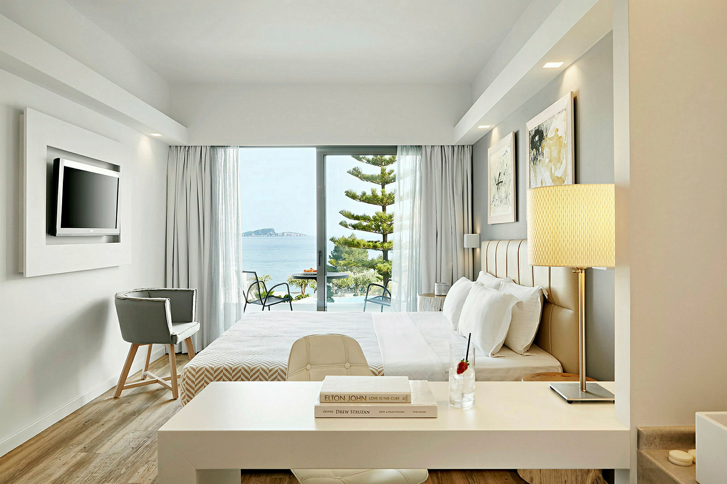 Ett hotellrum med modernt inredning och fönster som vetter mot en balkong med utsikt över havet. (AI-genererad text)