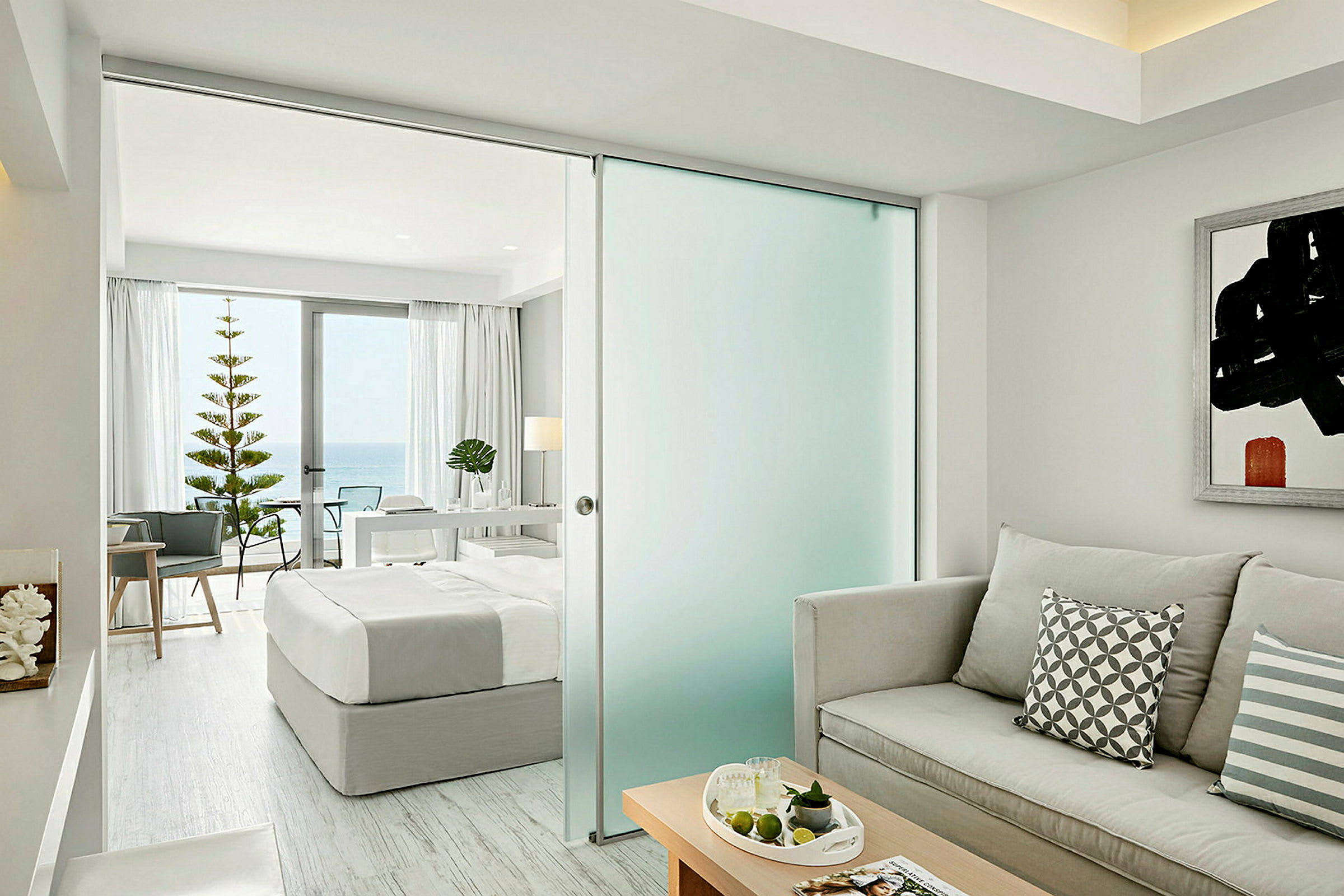 Ett ljust och modernt hotellrum med en säng, soffa och utsikt mot havet genom stora fönster. (AI-genererad text)