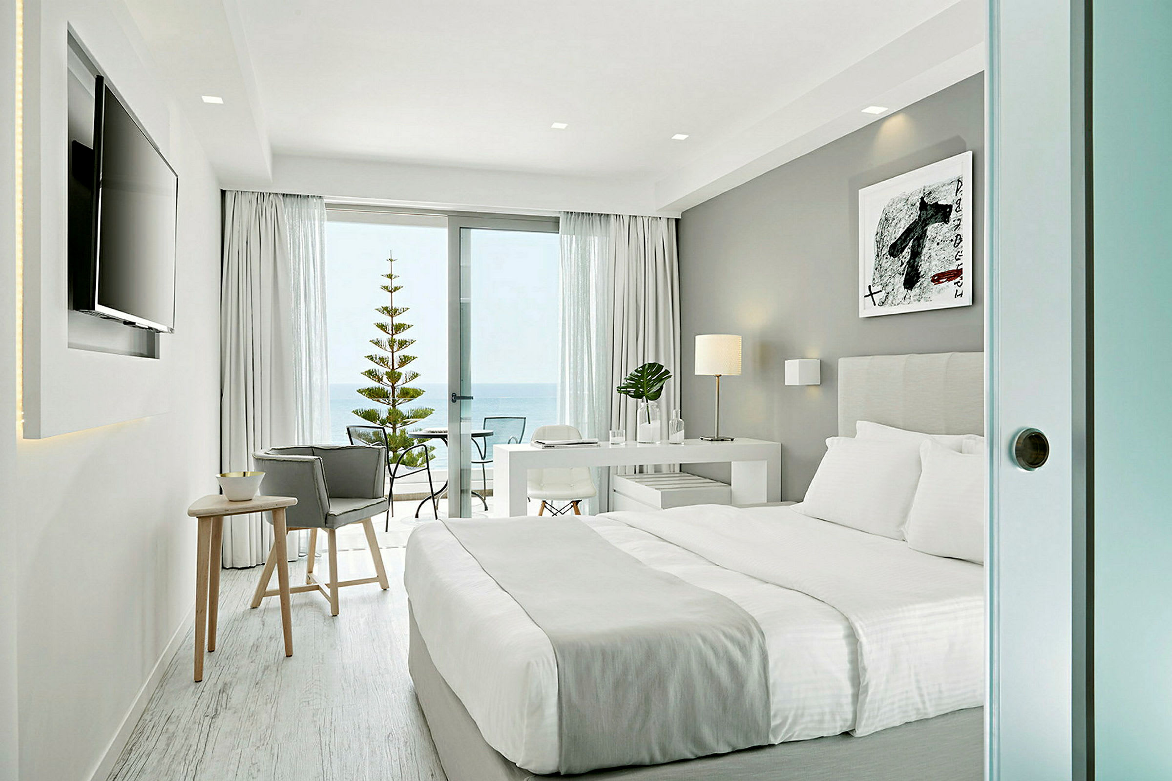 Modernt hotellrum med dubbelsäng, skrivbord och skjutdörr ut till en balkong med havsutsikt. (AI-genererad text)